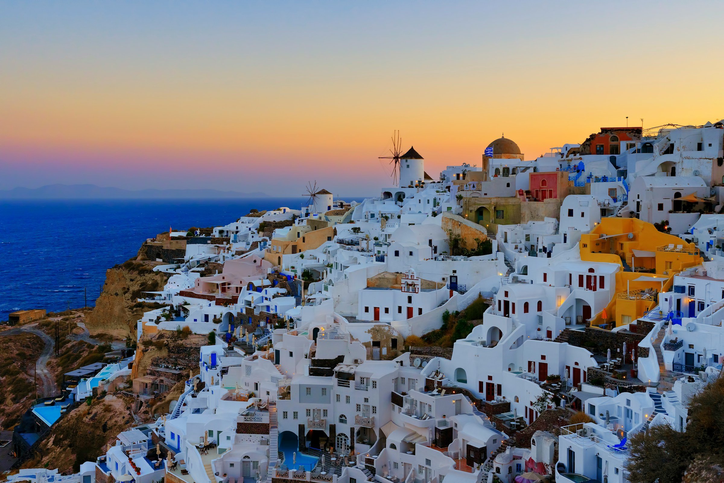 Utsikt over Oía på Santorini, Hellas, ved solnedgang. Hvite og fargerike bygninger med en vindmølle i bakgrunnen, mot en himmel som skifter fra oransje til blått