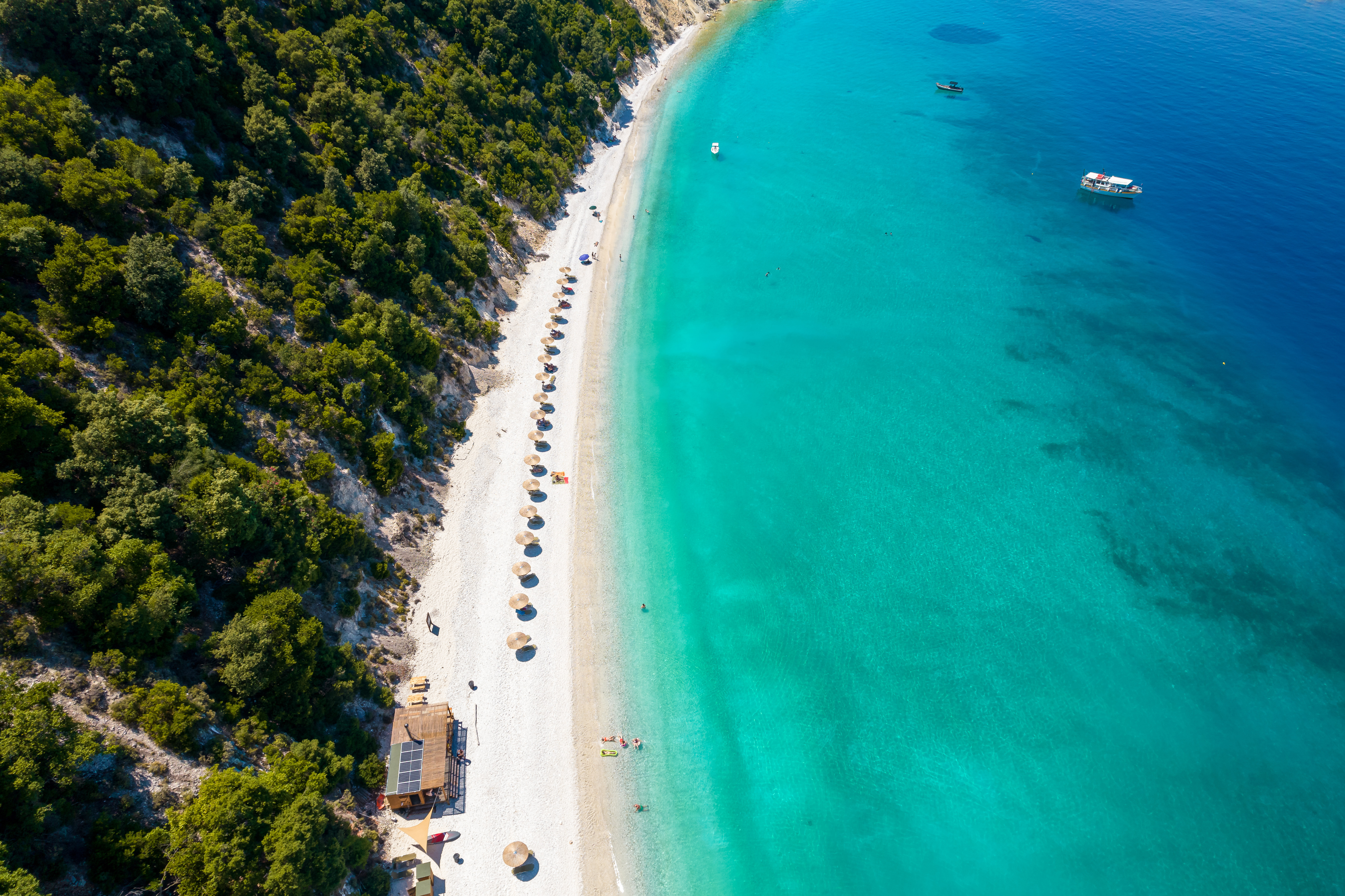 Flyfoto av en vakker strand med turkisblått vann, omgitt av frodig grønn vegetasjon. Parasoller er jevnt fordelt langs den hvite sandstranden. En båt ligger til rette på det rolige havet