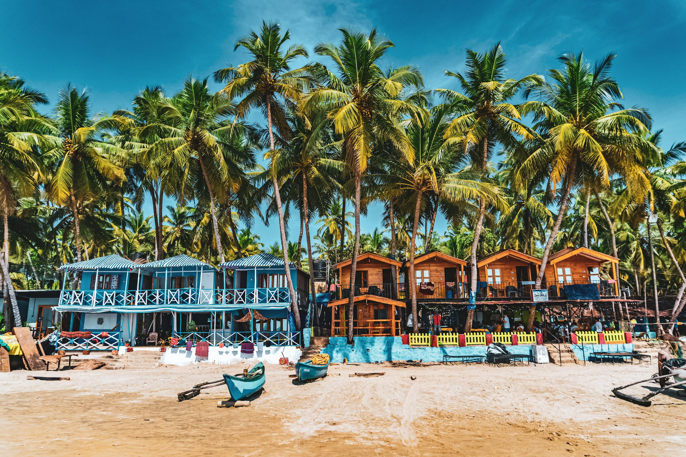 Colorful beach huts under tall palm trees on a sunny tropical beach, showcasing vibrant seaside architecture.