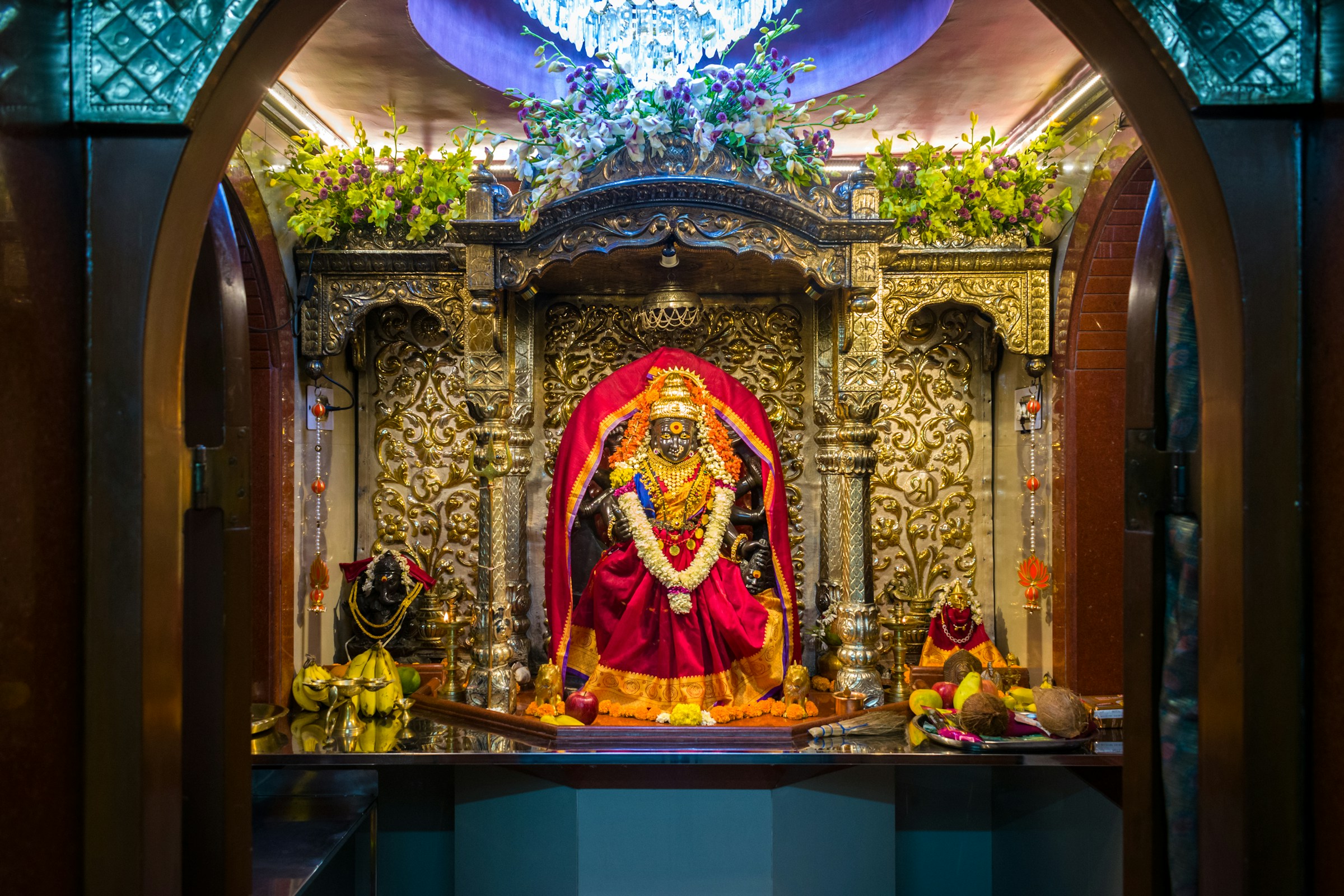 Intricately decorated Hindu temple altar with a deity statue adorned in red garments and garlands, surrounded by ornate silver carvings and floral arrangements.