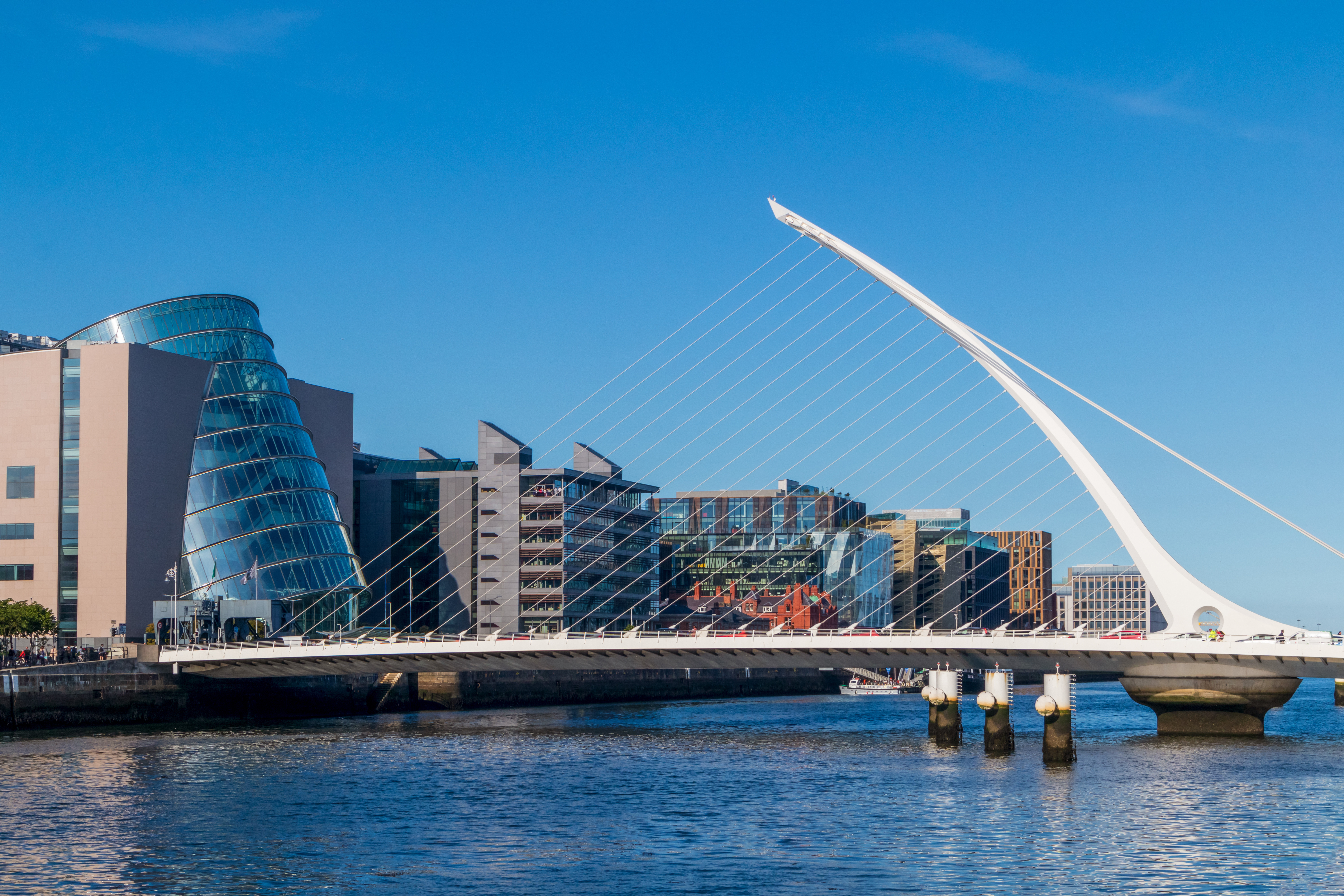 Travel to Dublin - Samuel Beckett Bridge in against a clear blue sky, with modern buildings and water reflections.