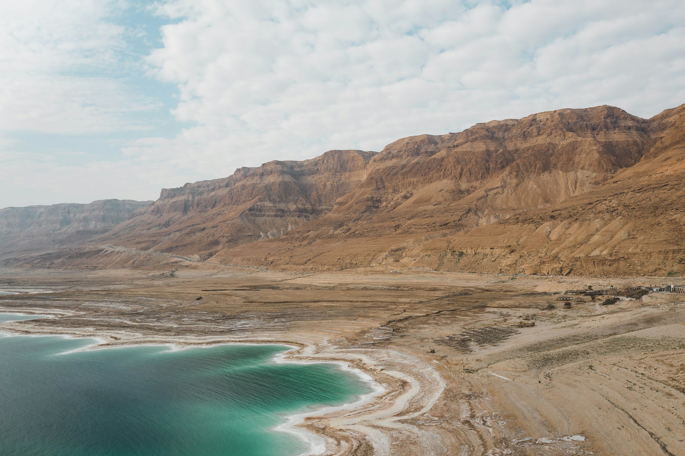 Aerial view of the Dead Sea coastline with rugged hills and a partly cloudy sky in the background, showcasing the unique natural landscape.