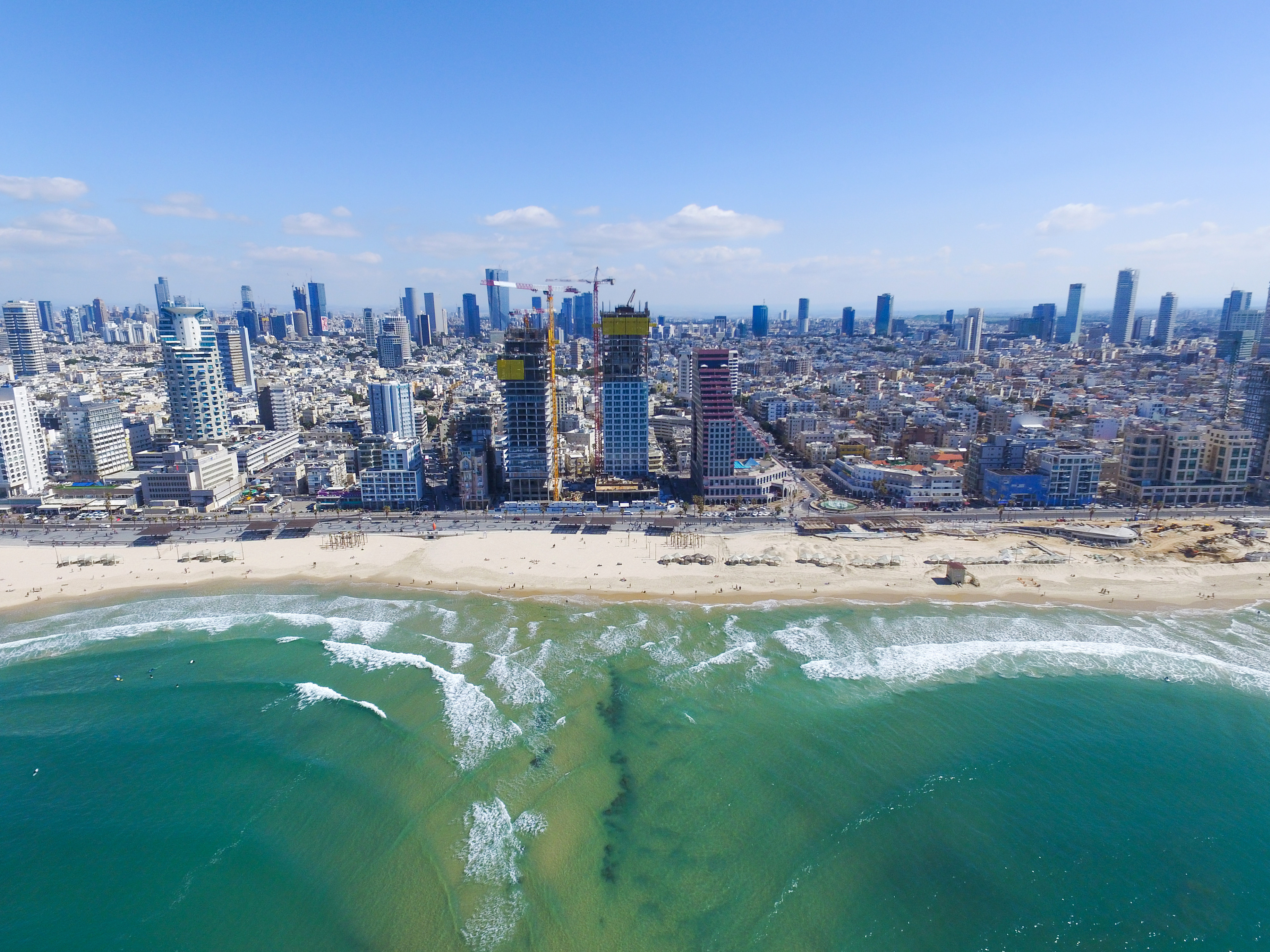 Luftfoto av kystlinjen i Tel Aviv med høye skyskrapere, byggeplasser og bølger som slår mot stranden under en klar blå himmel