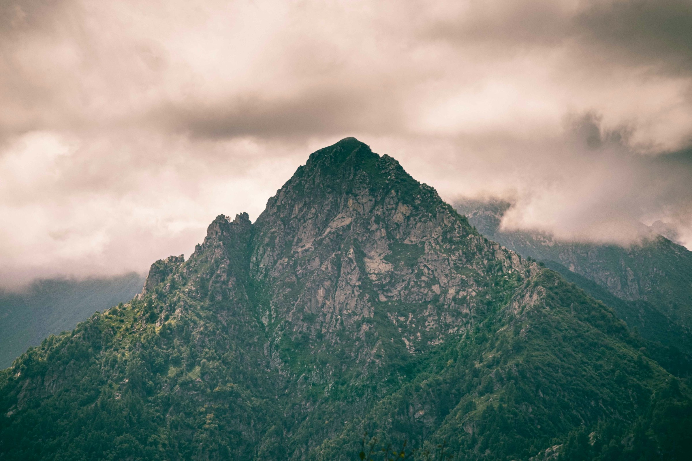 Mountain peak with steep cliffs and lush greenery under a dramatic cloud-covered sky in Alagna, Italy.