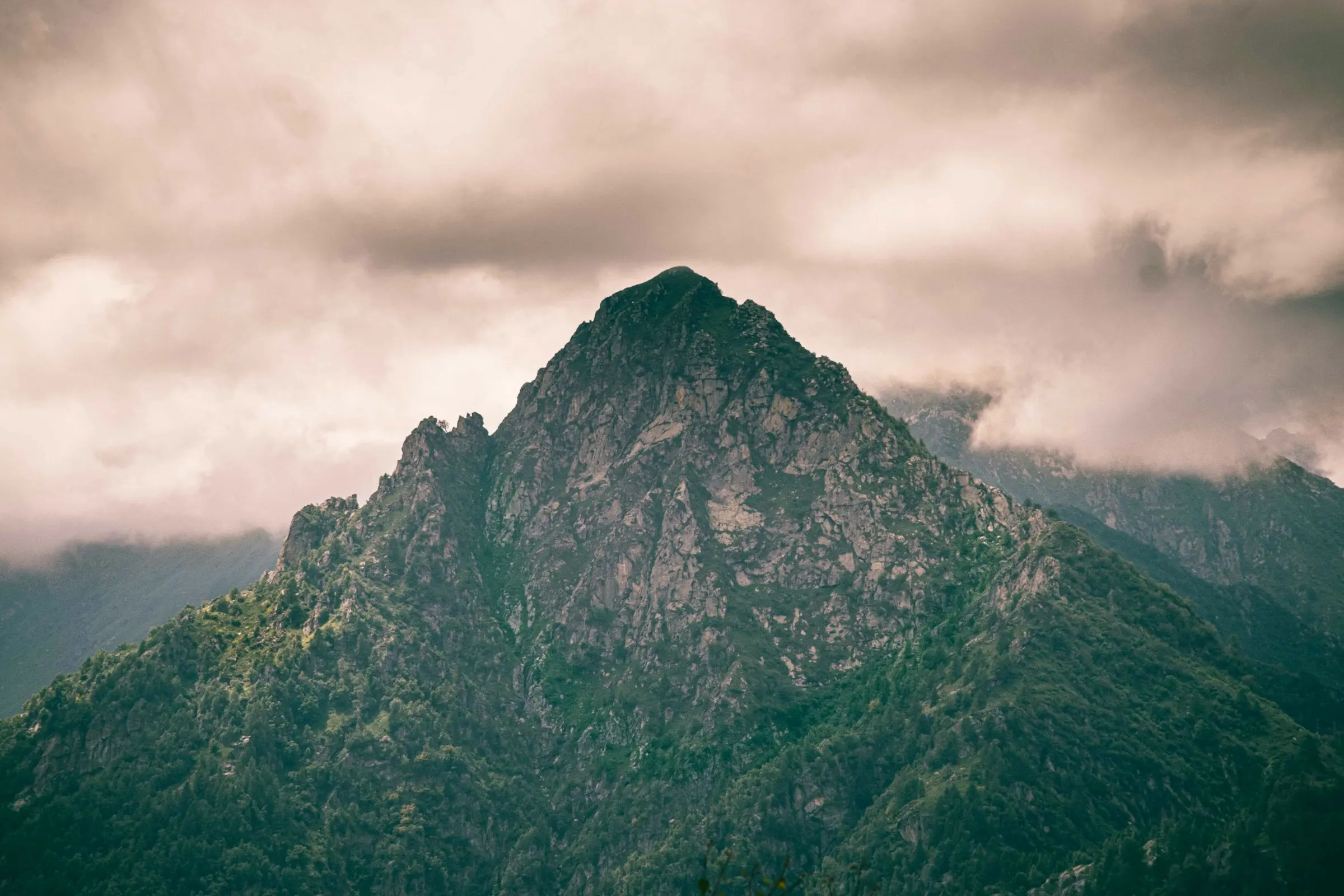 Mountain peak with steep cliffs and lush greenery under a dramatic cloud-covered sky in Alagna, Italy.