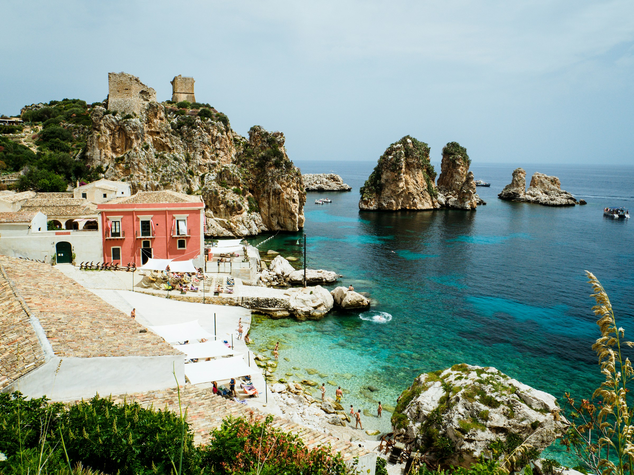 Scenic view of Tonnara di Scopello, Sicily, Italy, showcasing crystal-clear turquoise waters, rugged rock formations, historic buildings.