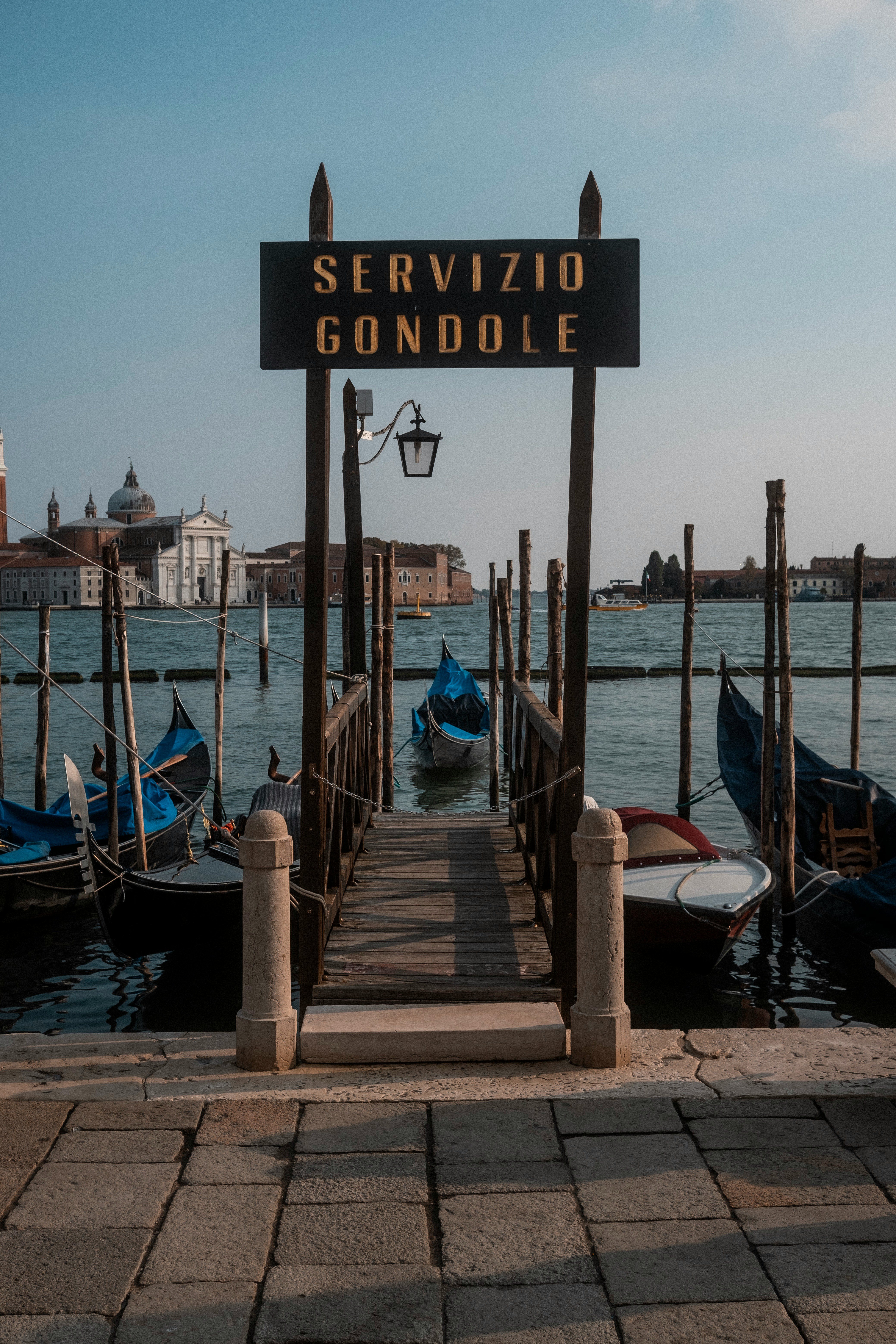 Gangvei til gondolterminal i Venezia med skilt merket "Servizio Gondole", omgitt av gondoler og vann med venetianske bygninger i bakgrunnen