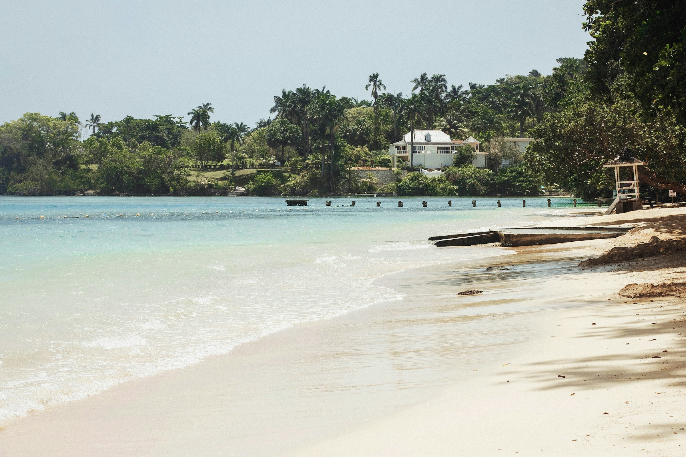 Tropical beach with clear turquoise water, sandy shoreline, and lush greenery, featuring a white house set back from the coastline.