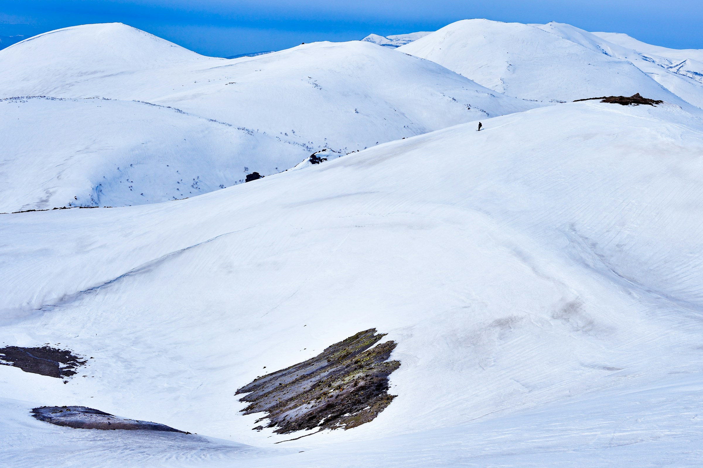Snødekte fjelltopper under en klar blå himmel, med spor av fjellformasjoner og en ensom turgåer i Niseko, Japan.