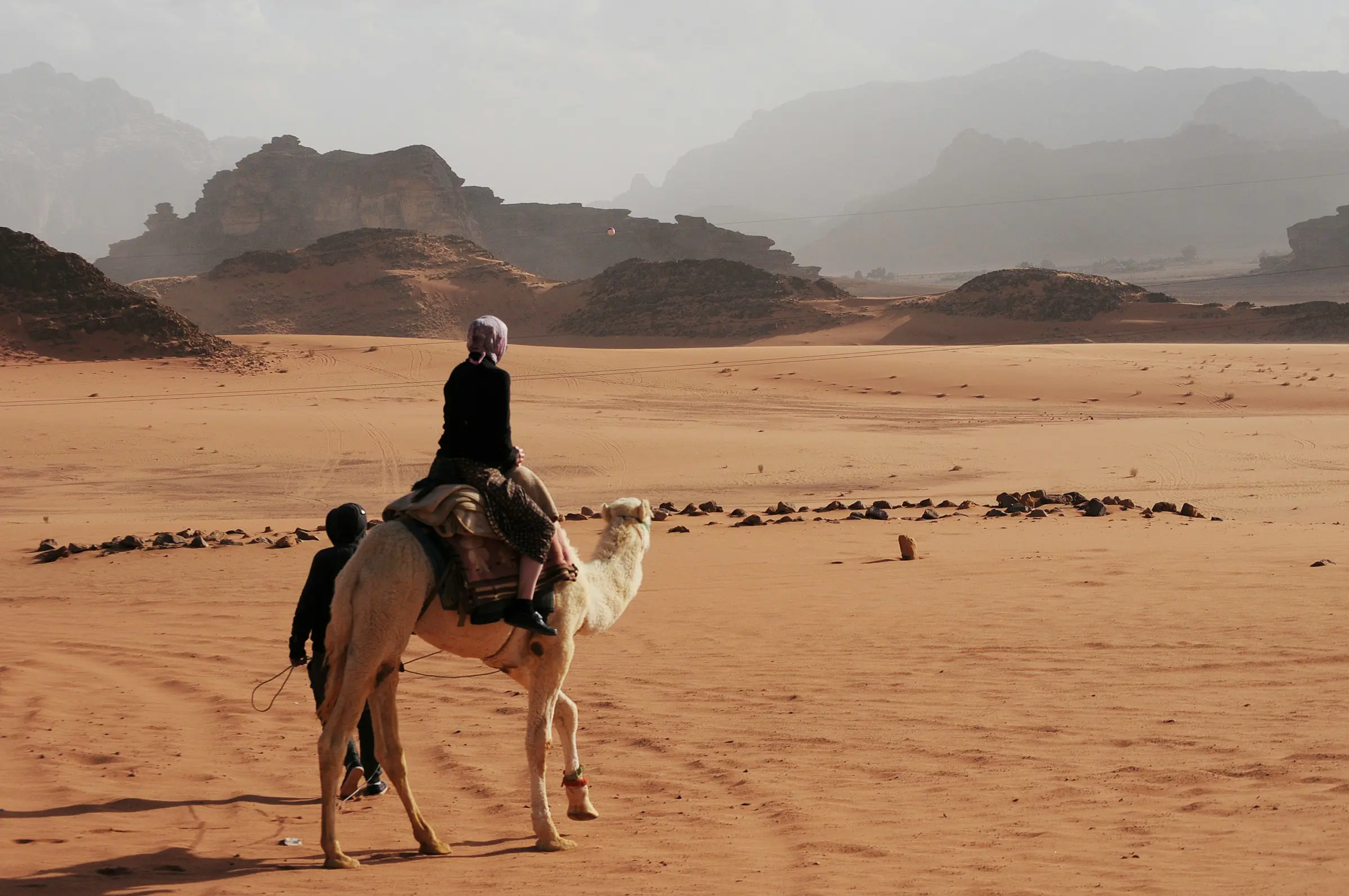 Person riding a camel in the desert with rocky hills in the background, under a clear sky.
