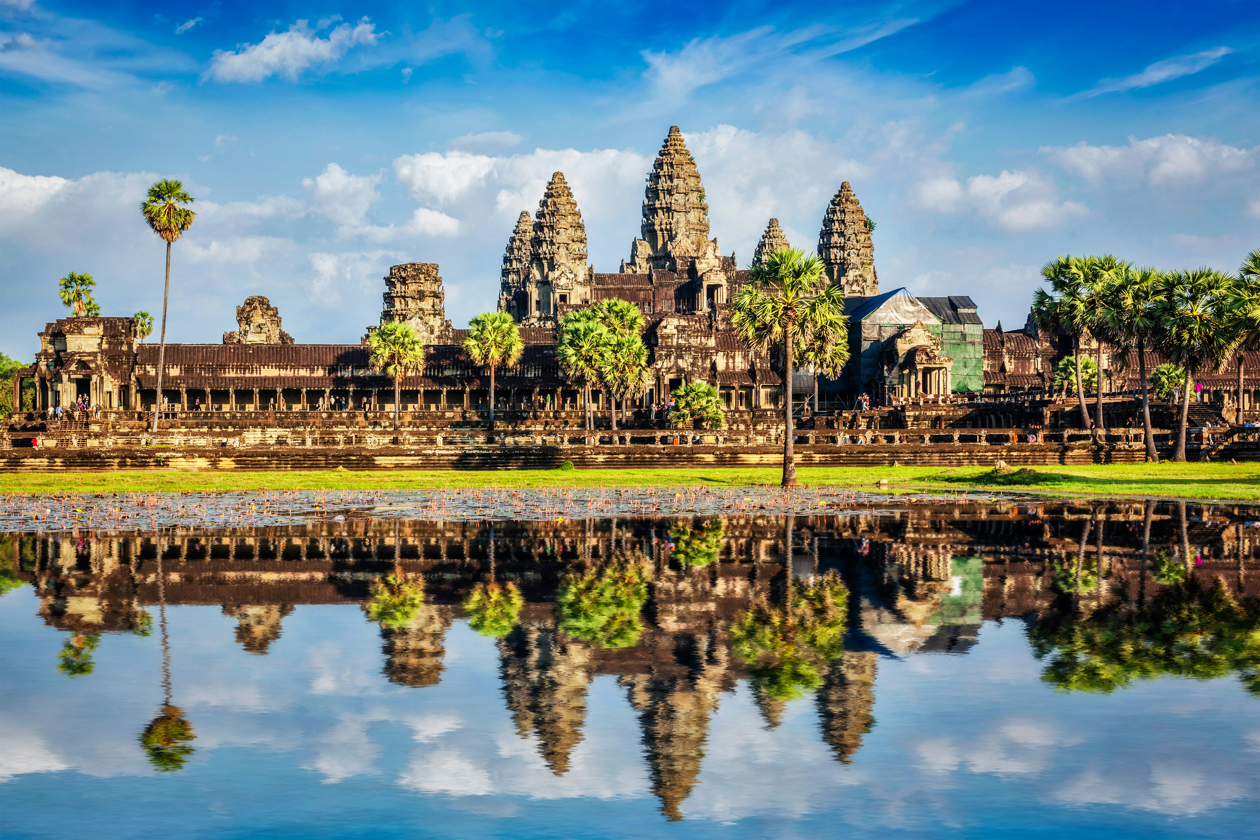 Ancient temple of Angkor Wat in Cambodia with clear reflection in a calm lake, surrounded by lush greenery under a bright blue sky.