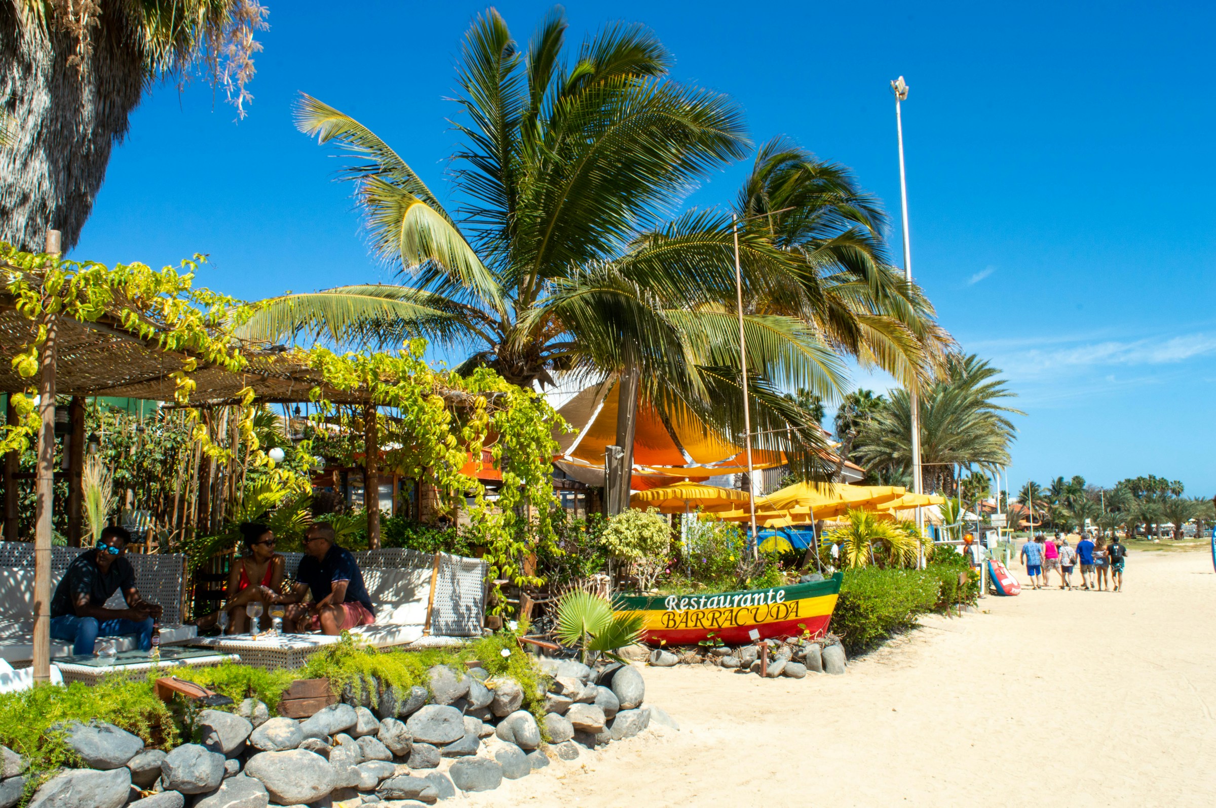 Beachside restaurant with palm trees and people enjoying a sunny day in a tropical setting.