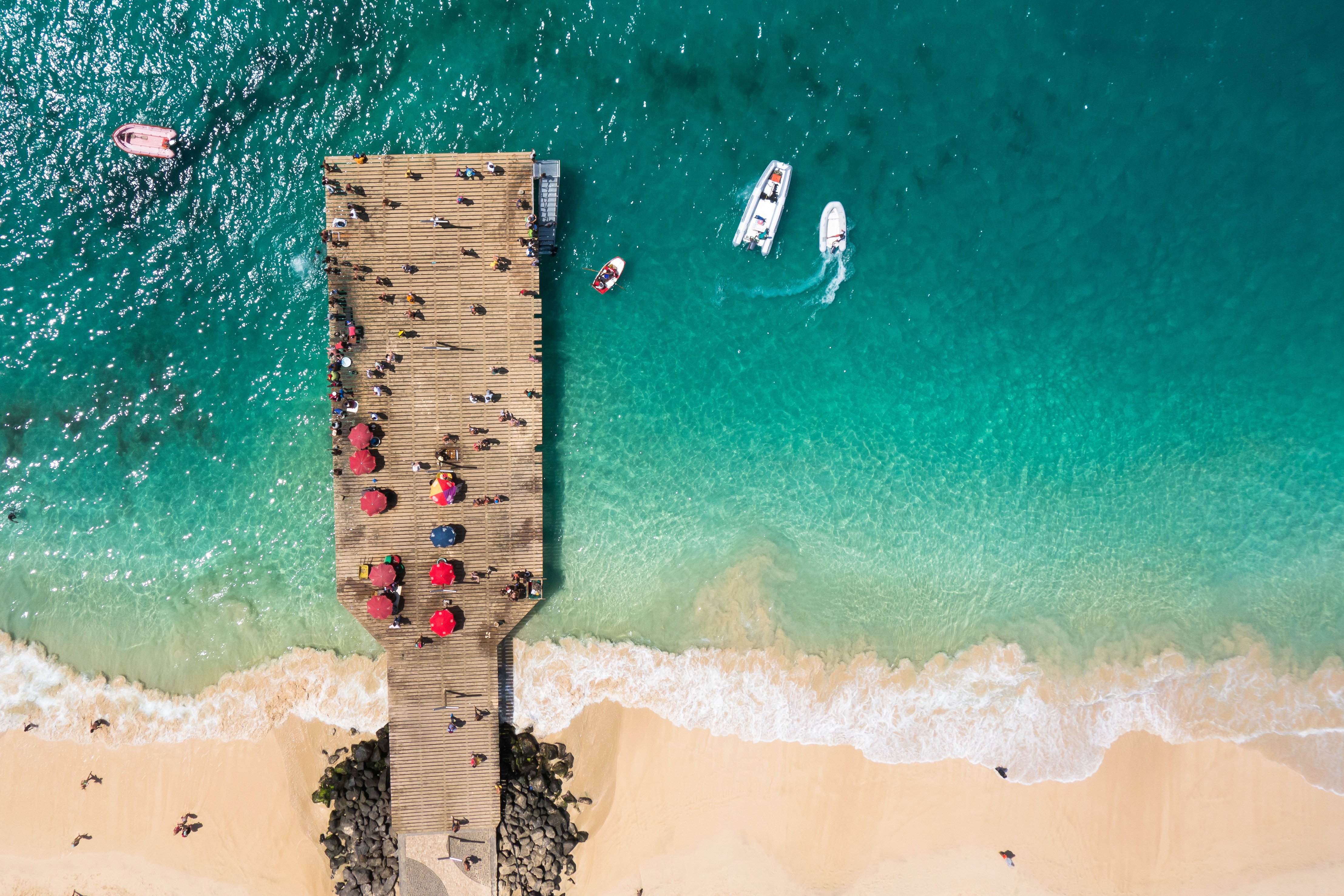 Travel to Sal - Aerial view of a wooden pier with people and red umbrellas overlooking turquoise ocean waters and sandy beach.
