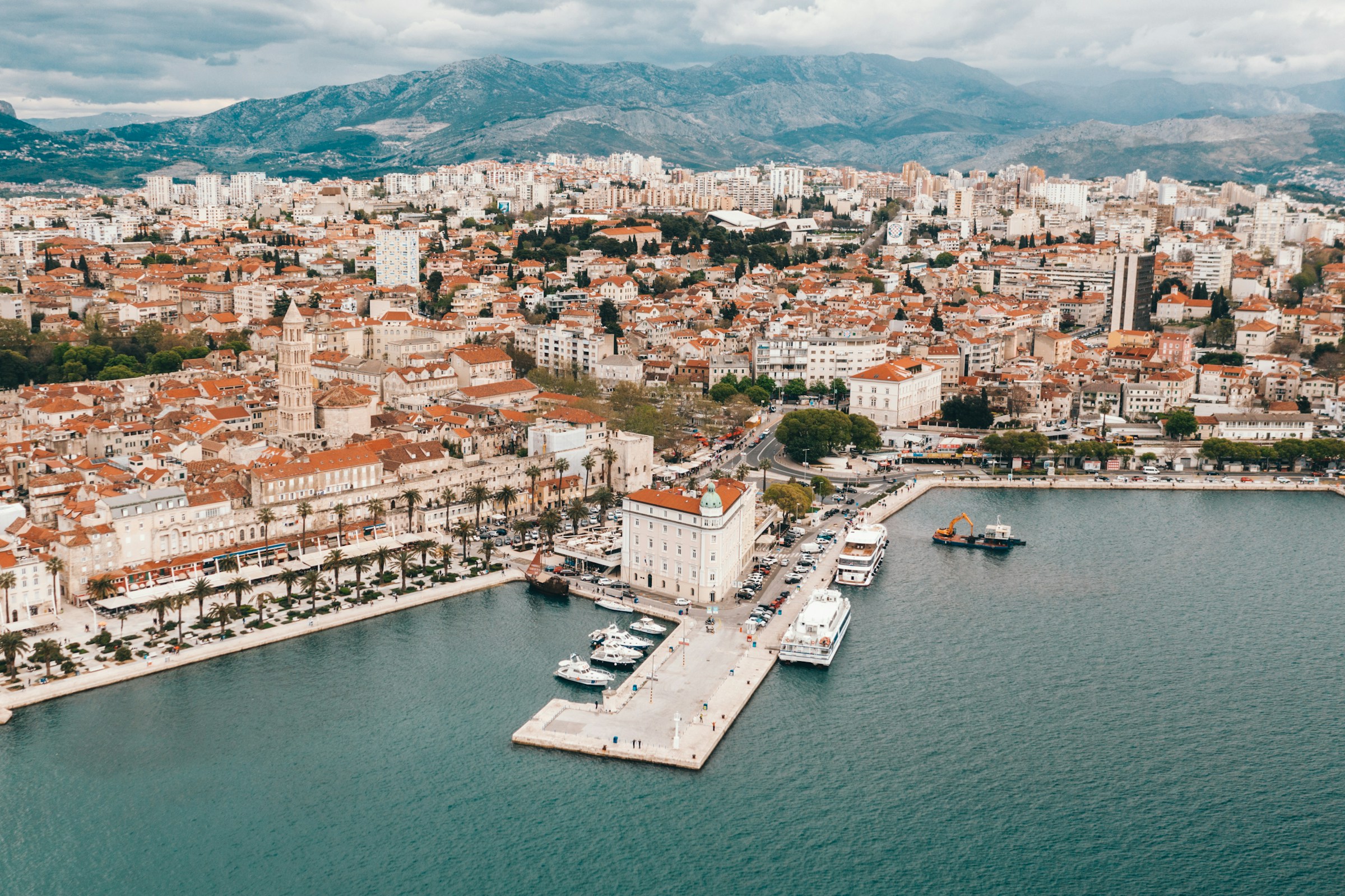 Aerial view of Split, Croatia, showcasing the historic cityscape with red-roofed buildings, waterfront promenade, and bustling harbor against a backdrop of mountains and cloudy skies