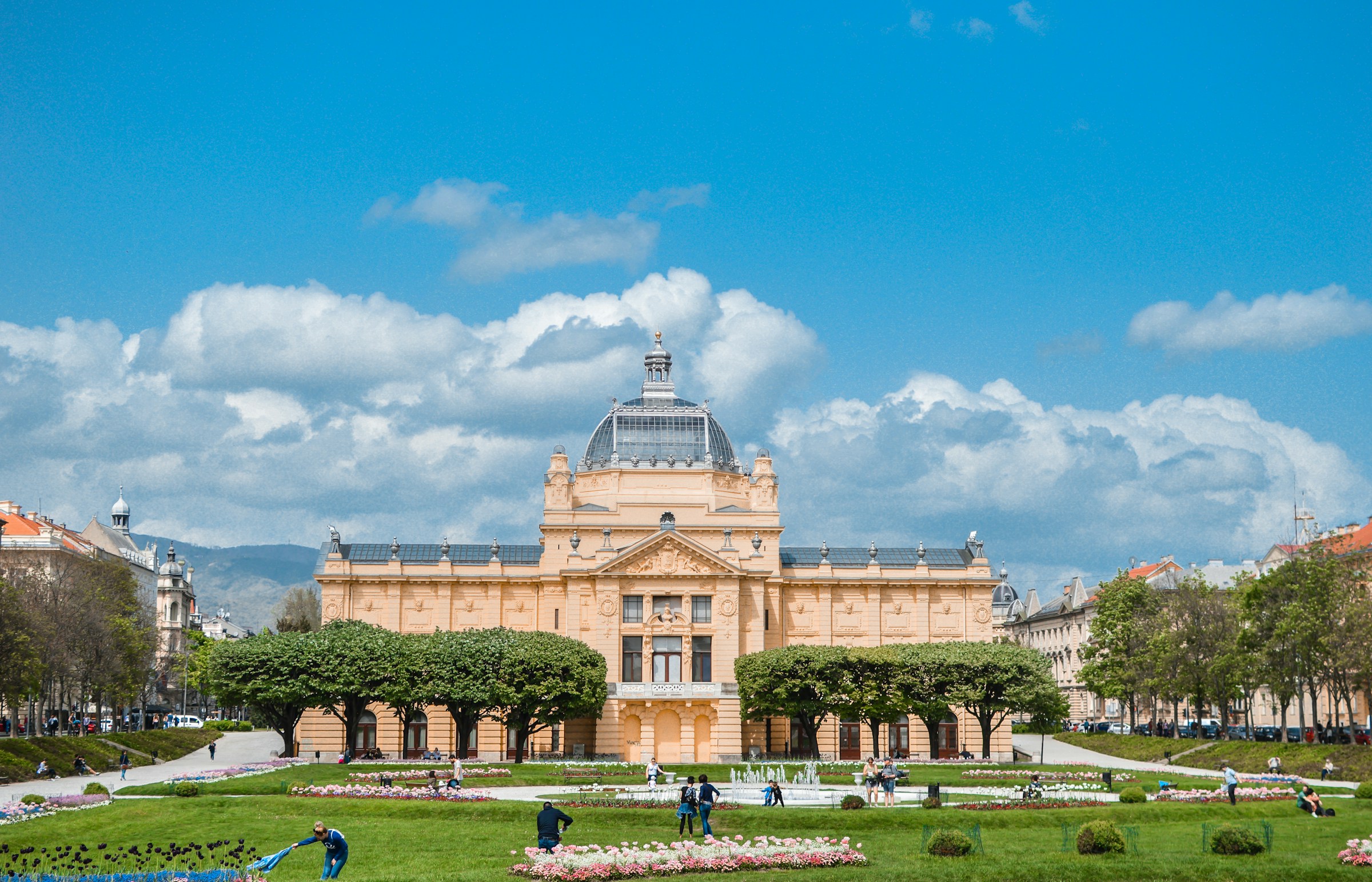 Historic Art Pavilion in Zagreb, Croatia under a blue sky with clouds, surrounded by green lawn and vibrant flowers