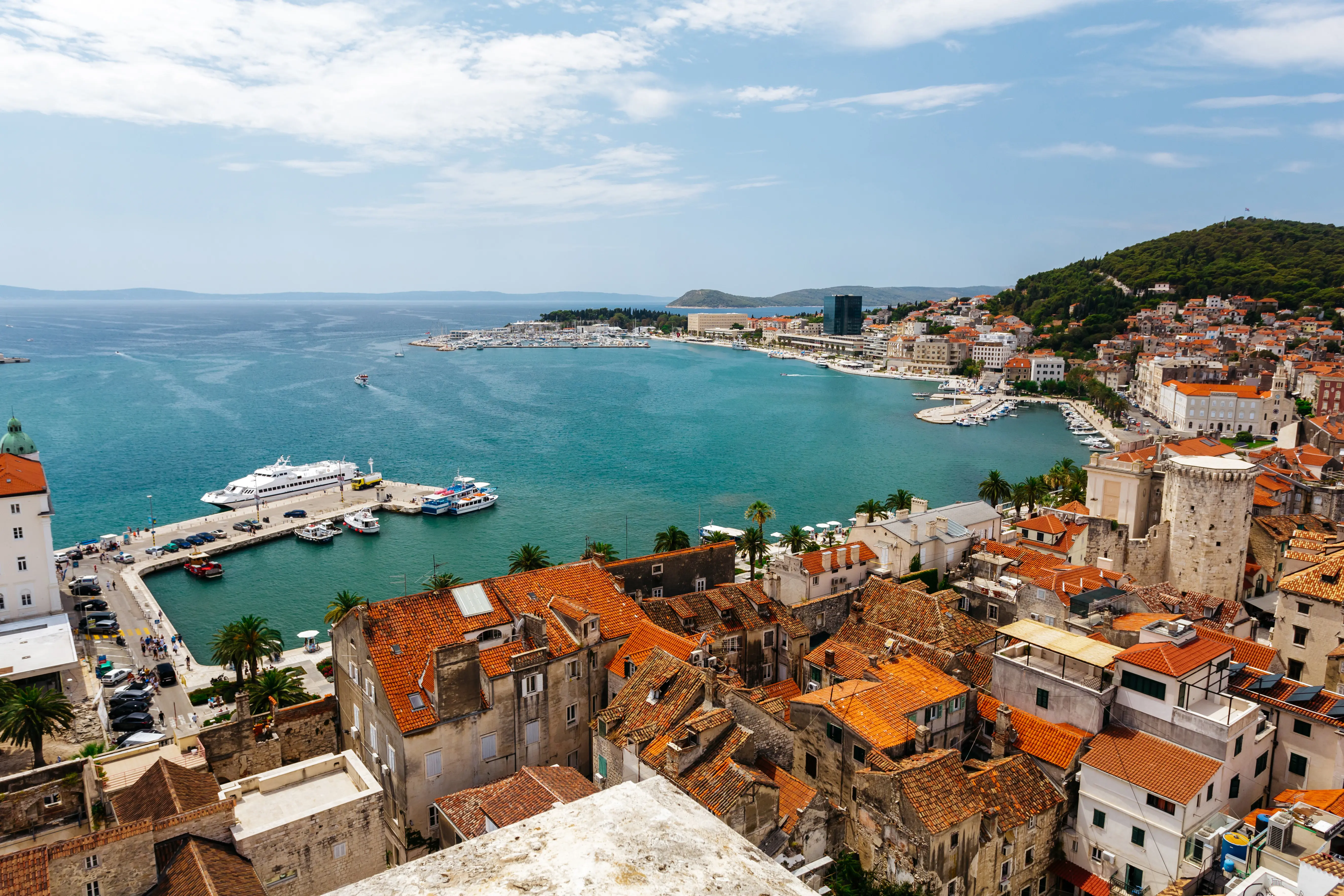 Travel to Split - Harbor view with red-roofed buildings, turquoise sea, boats docked, and lush green hillside.