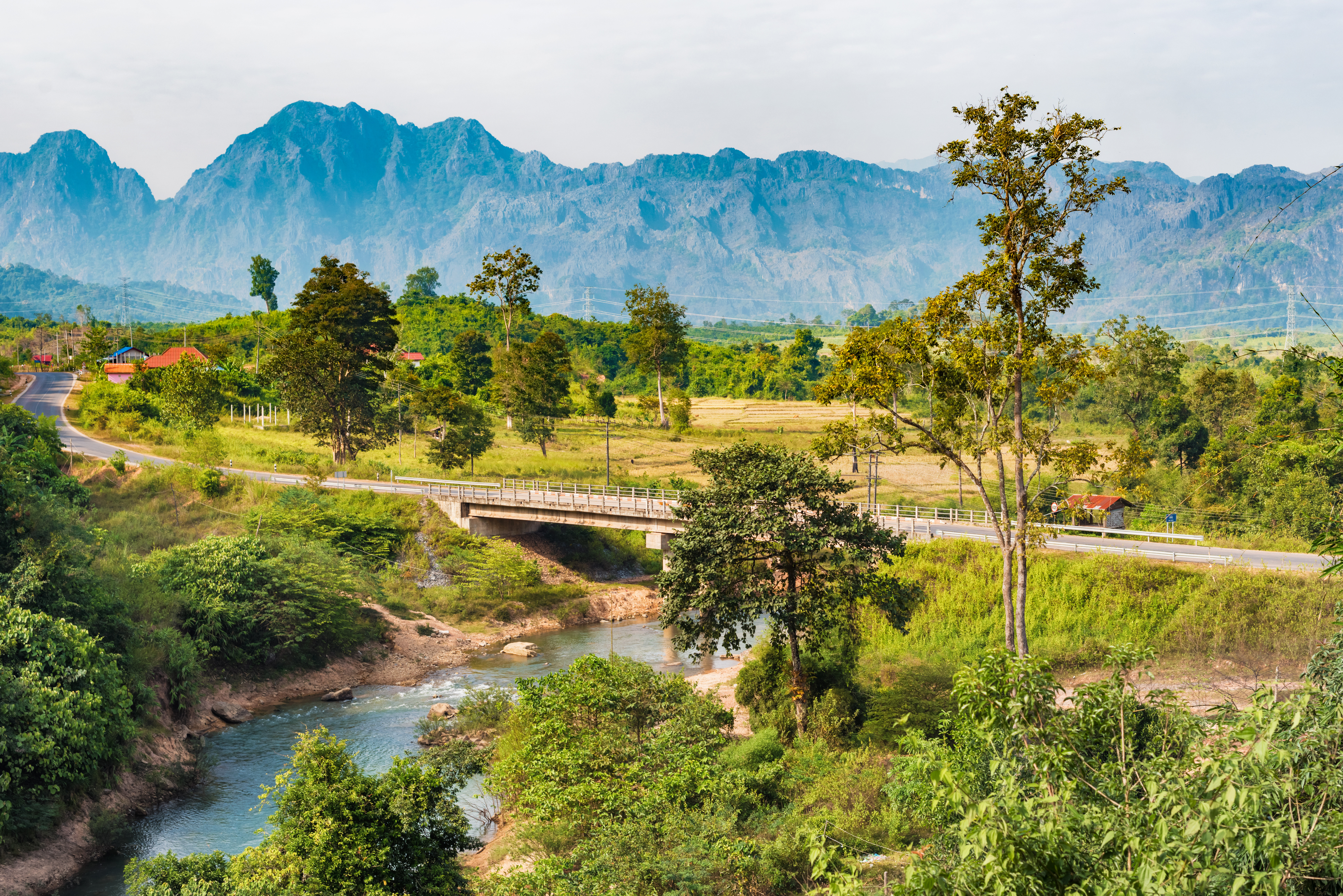 Scenic landscape with a river and bridge surrounded by lush greenery and mountains in the background under a clear sky