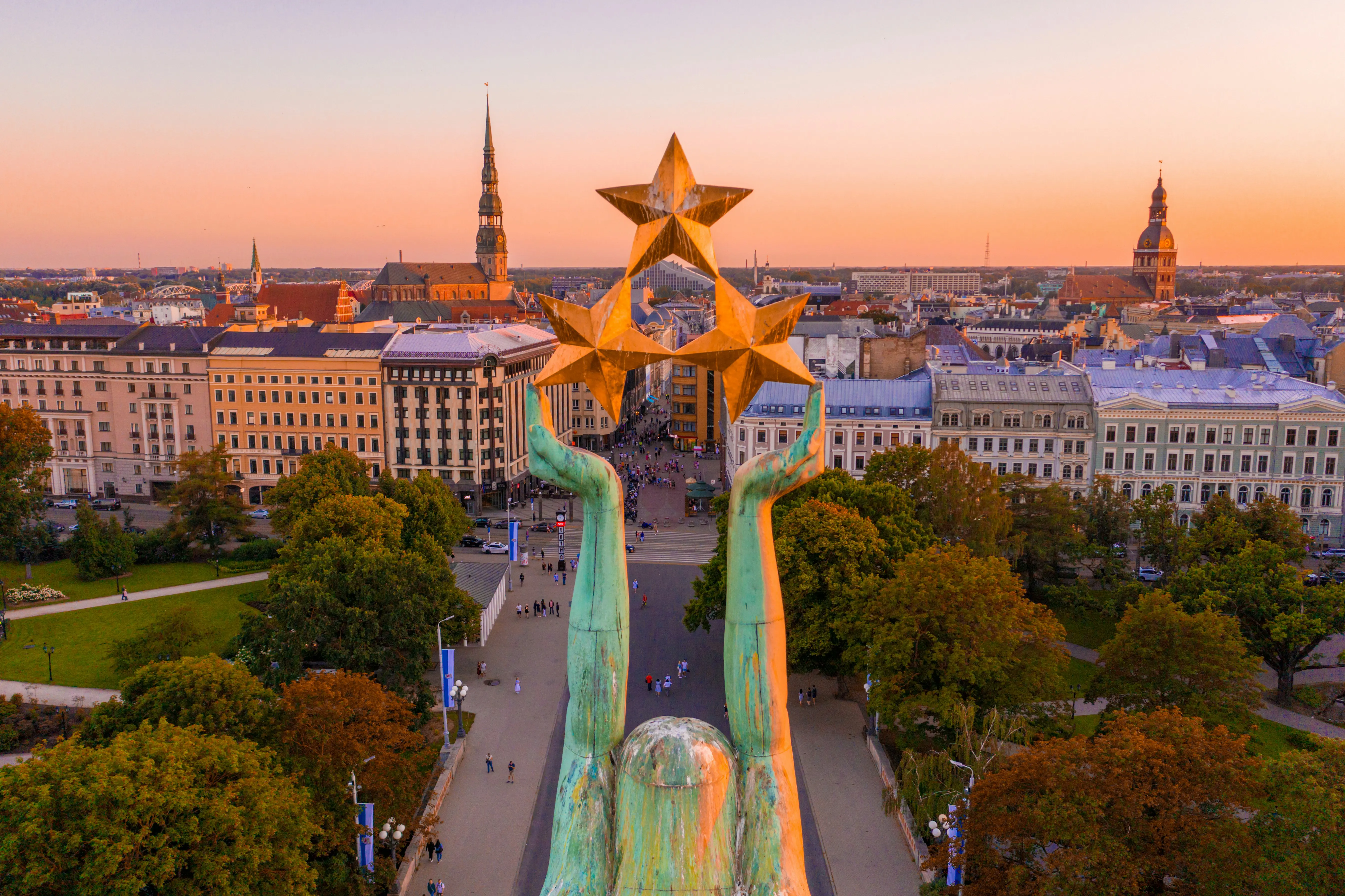Flyfoto av Frihetsmonumentet i Riga, Latvia, med gyldne stjerner på toppen av en søyle og byens silhuett i bakgrunnen ved solnedgang
