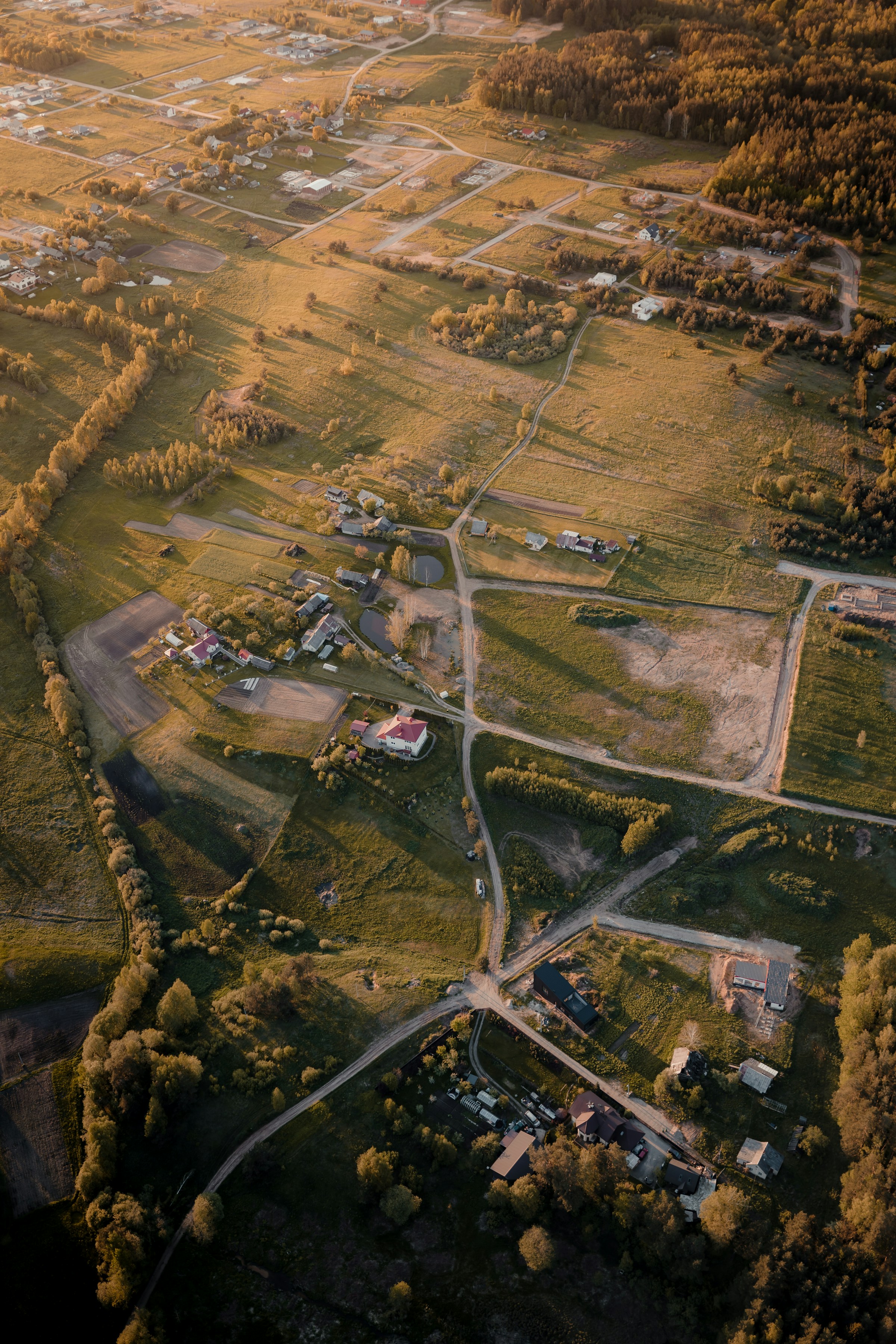 Aerial view of rural landscape during sunset, featuring sprawling fields, a few scattered houses, and intersecting dirt roads, with a backdrop of lush green forests