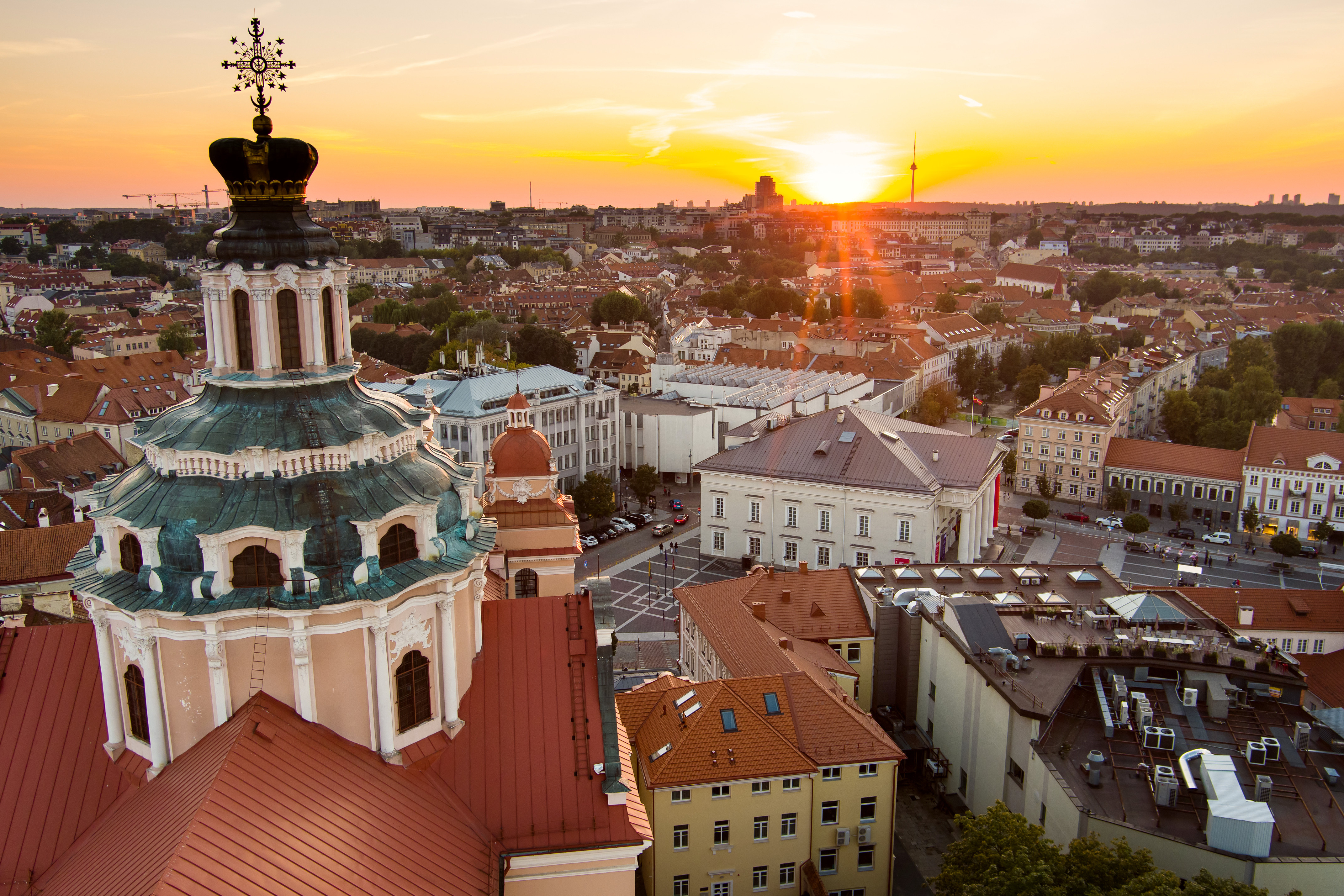Aerial view of Vilnius at sunset, featuring the Church of St. Catherine with its ornate architecture in the foreground, and the cityscape stretching into the horizon, bathed in golden light
