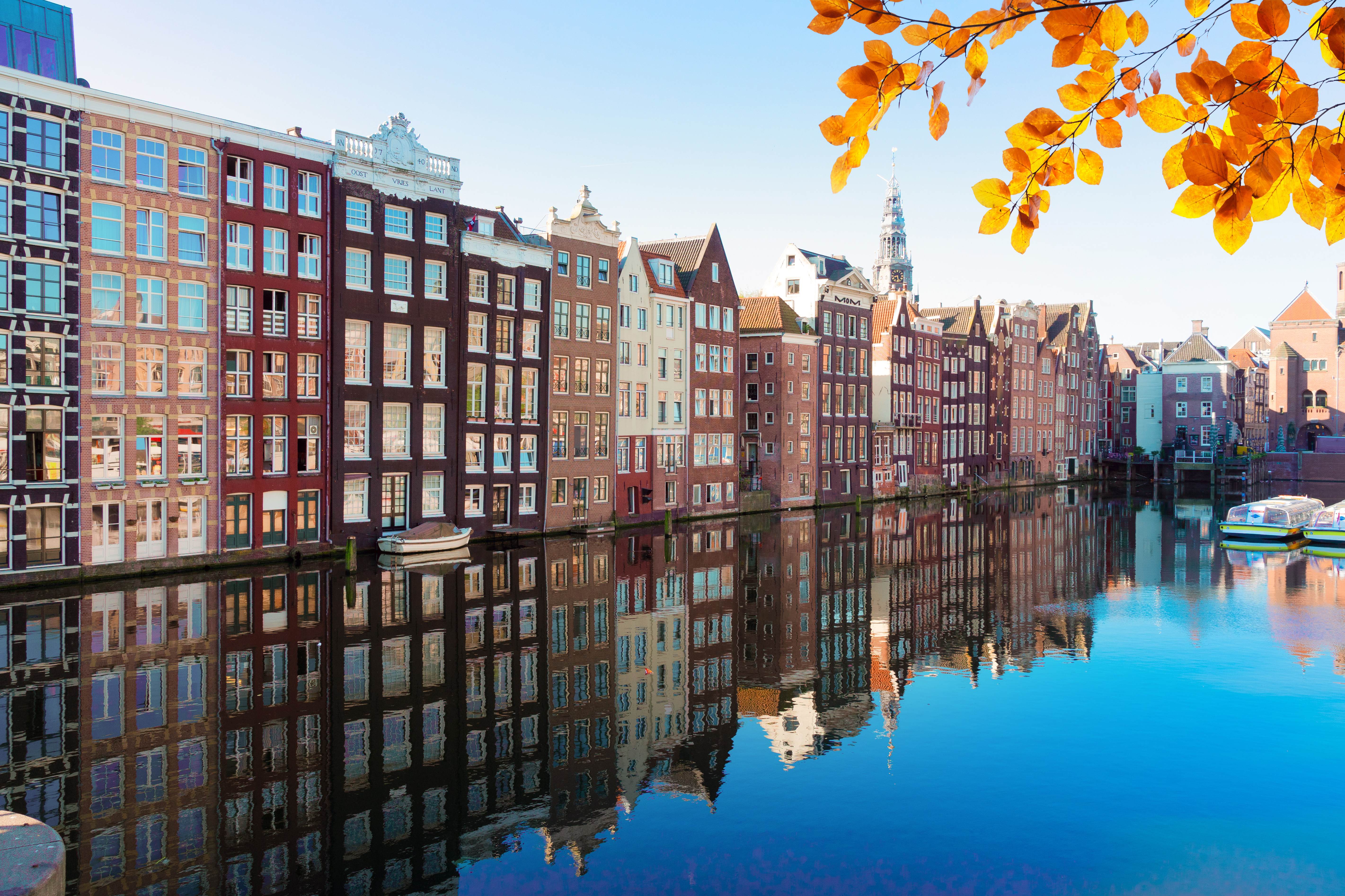 Husrekke langs en kanal i Amsterdam med fargerike blader i forgrunnen og speilbilde i vannet nedenfor, på en klar høstdag