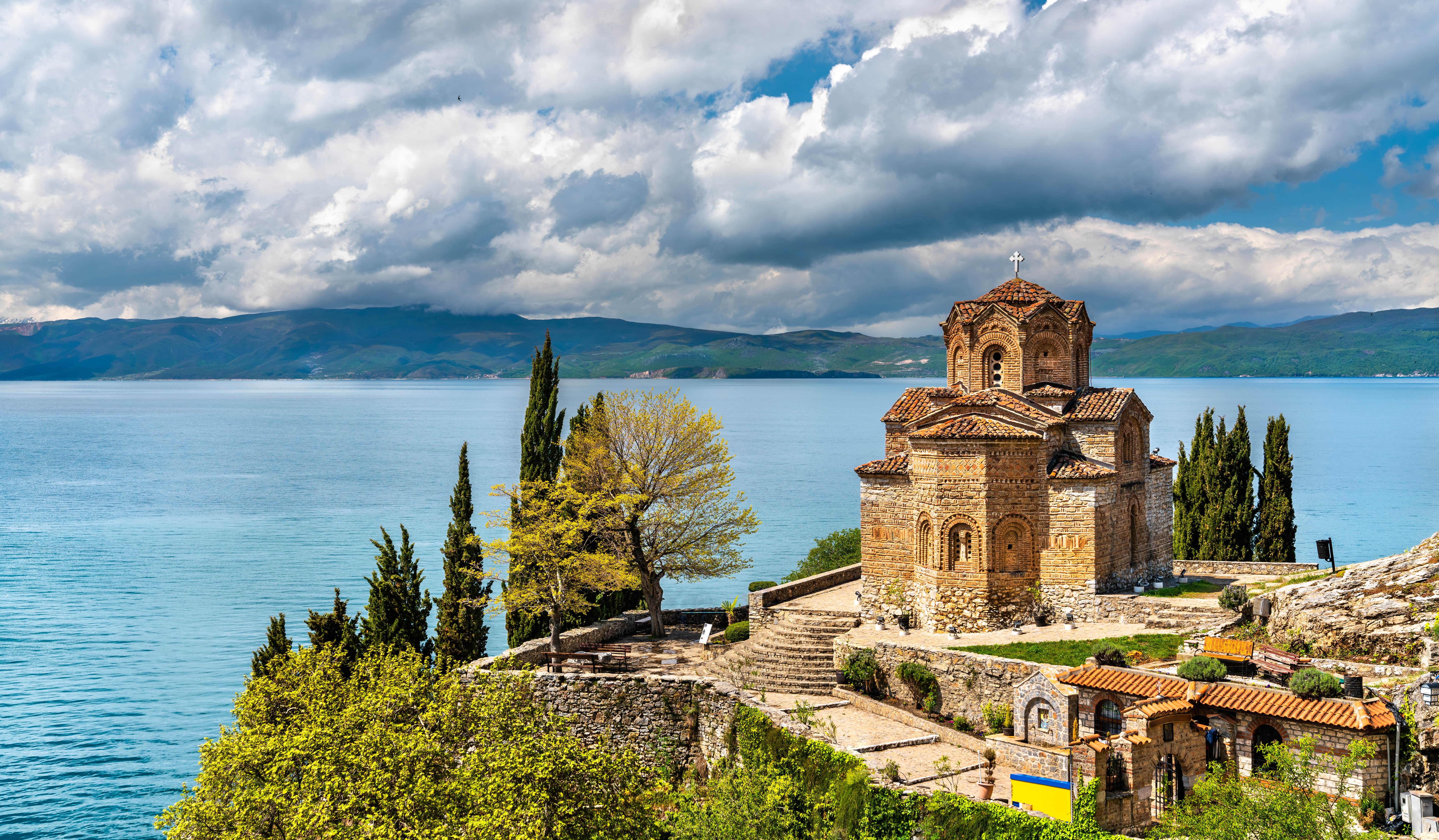 Reise til Nord-Makedonia - Historisk kirke ved bredden av en innsjø, omgitt av trær og grønne landskap, med dramatiske skyer i bakgrunnen.