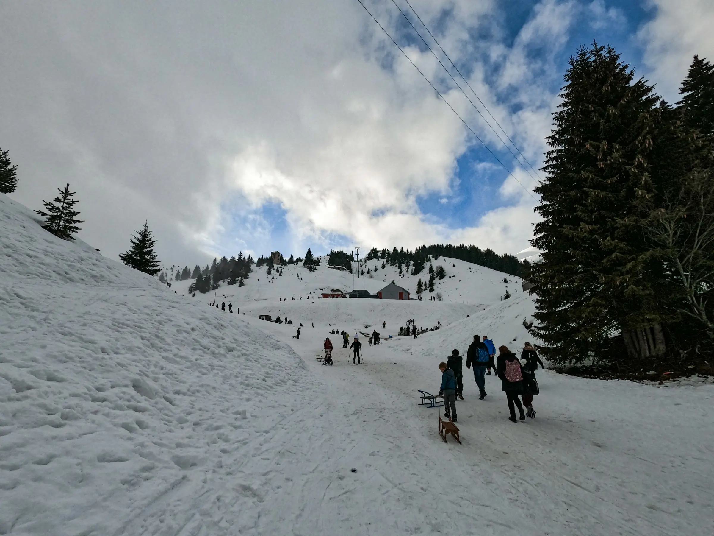 Folk går opp en snødekt skibakke med bakker og fjell og overskyet himmel i bakgrunnen