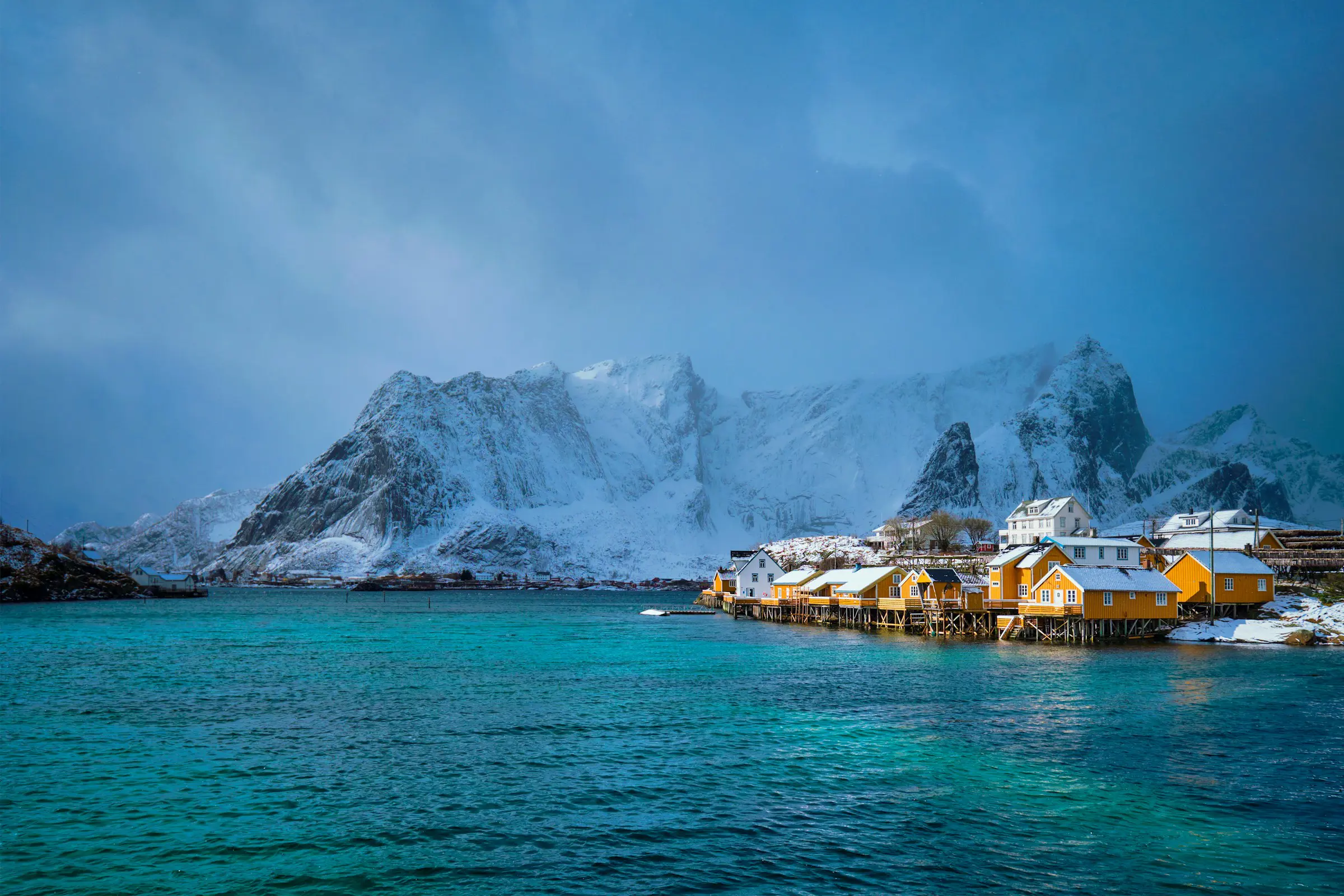 Snow-covered mountains and turquoise water surround traditional yellow cabins on stilts in Lofoten, Norway.