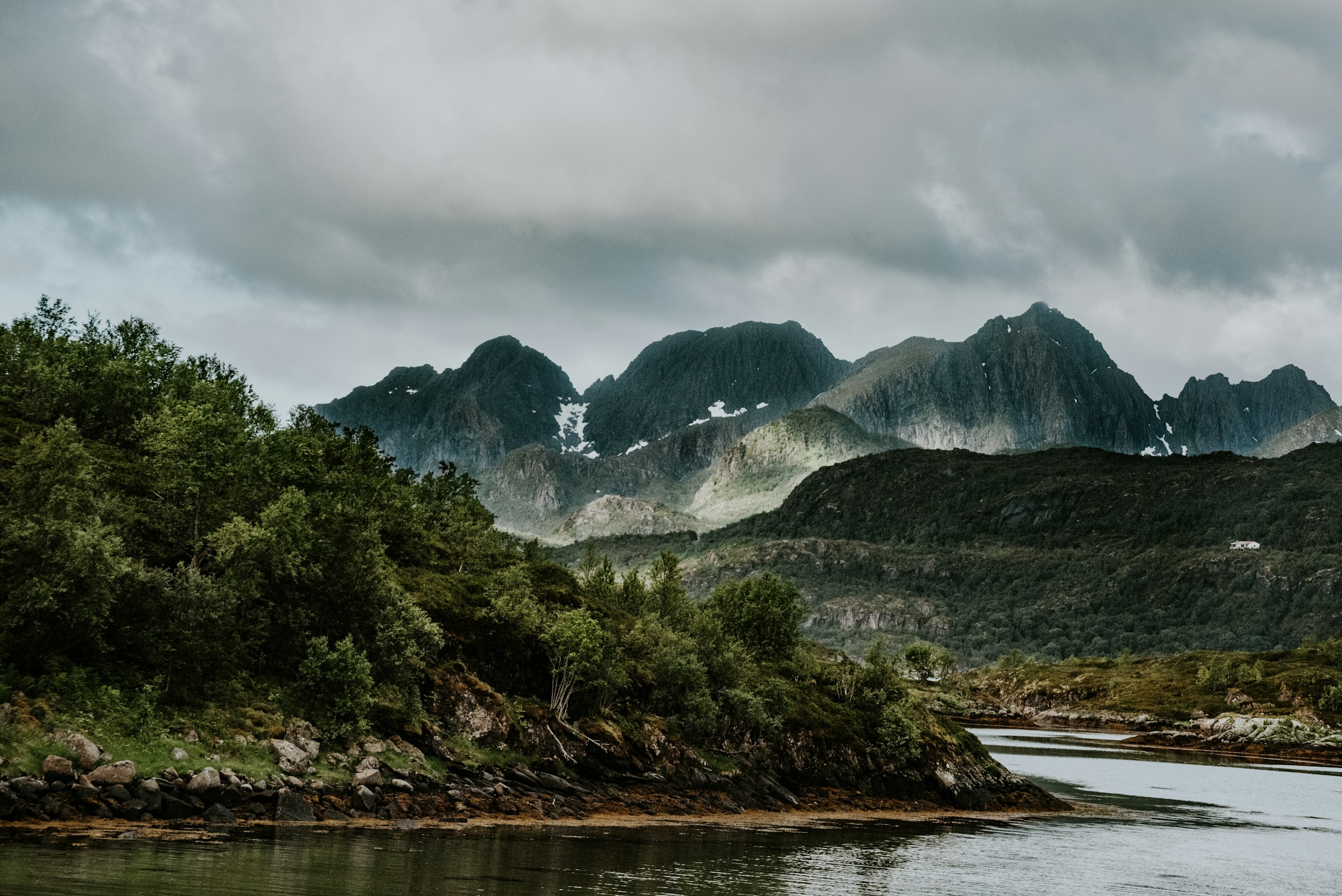 Mountain landscape with lush greenery and a calm river under a cloudy sky, showcasing rugged peaks in the distance.