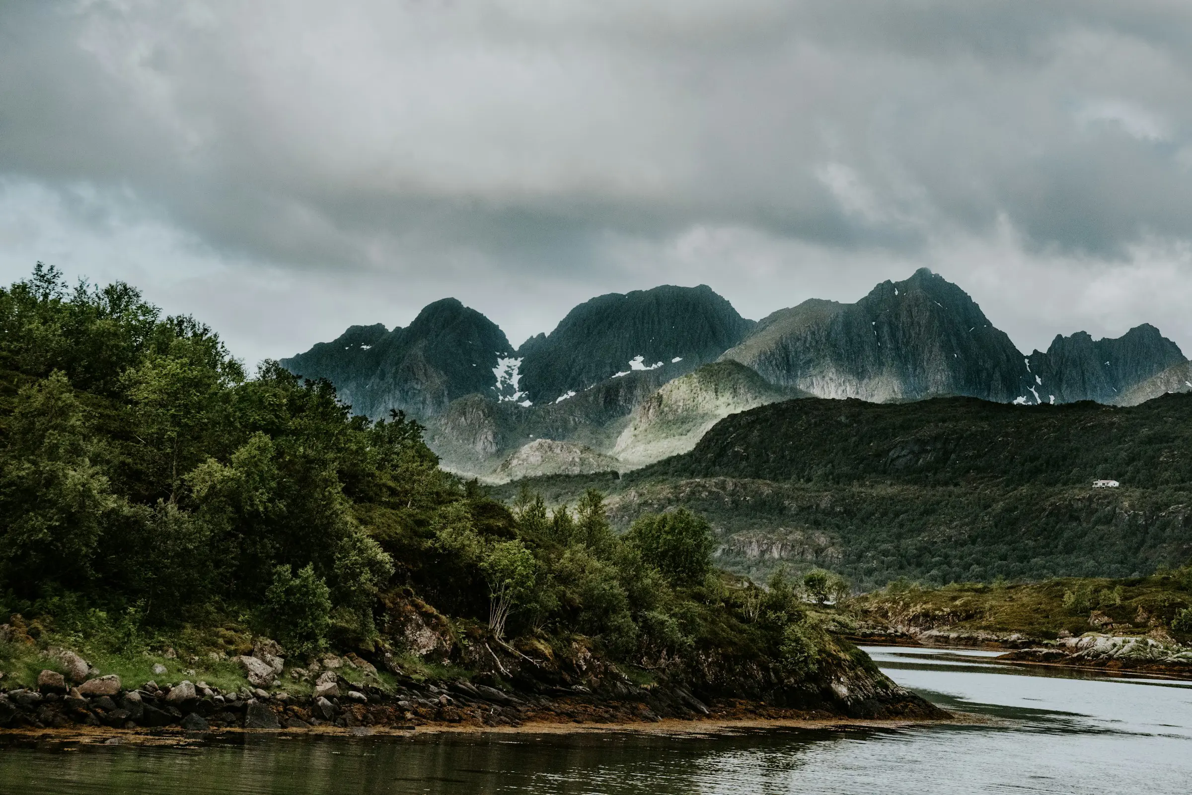 Mountain landscape with lush greenery and a calm river under a cloudy sky, showcasing rugged peaks in the distance.