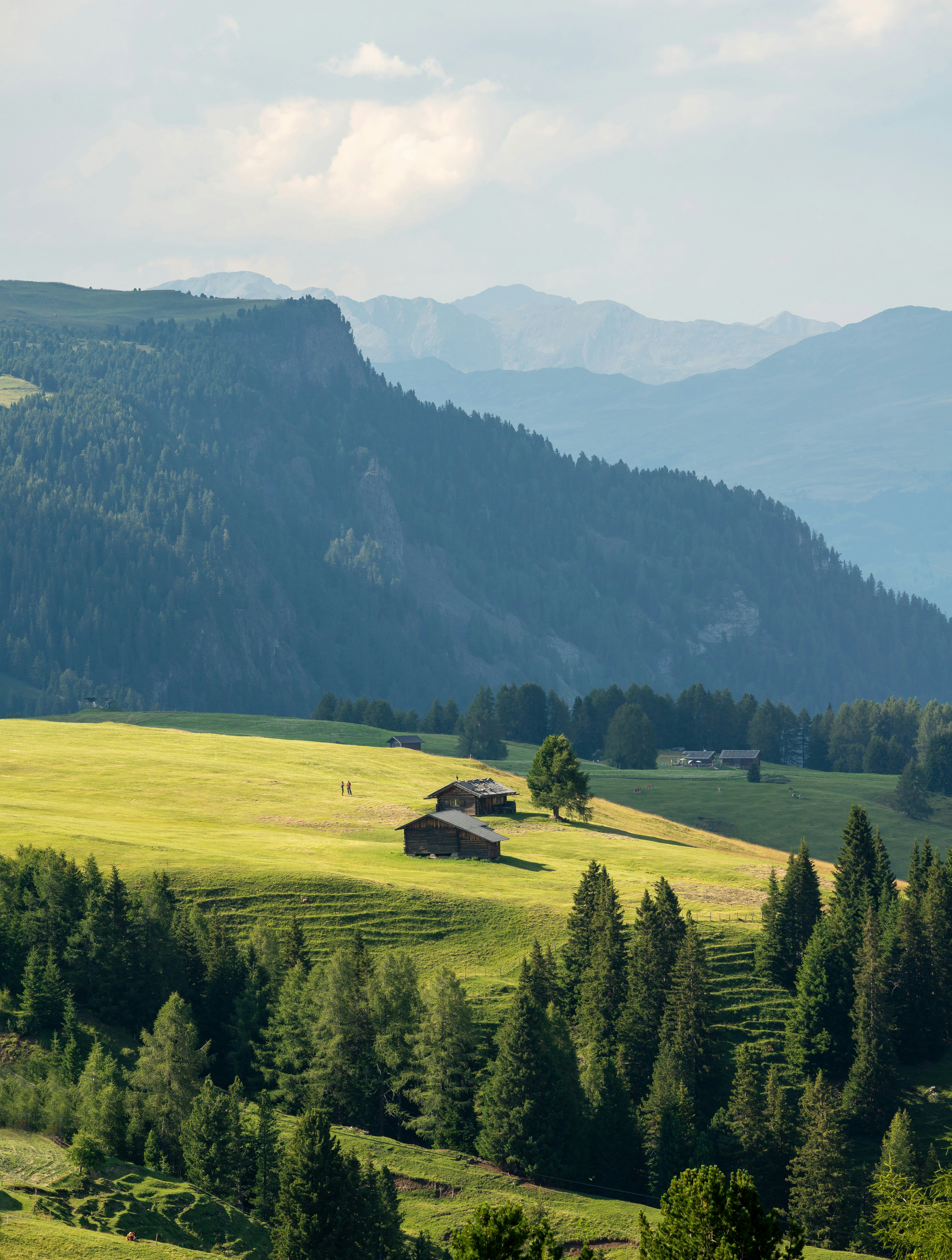 Mountain landscape featuring lush green hills, scattered trees, and rustic wooden cabins under a clear sky, with mountain peaks in the background