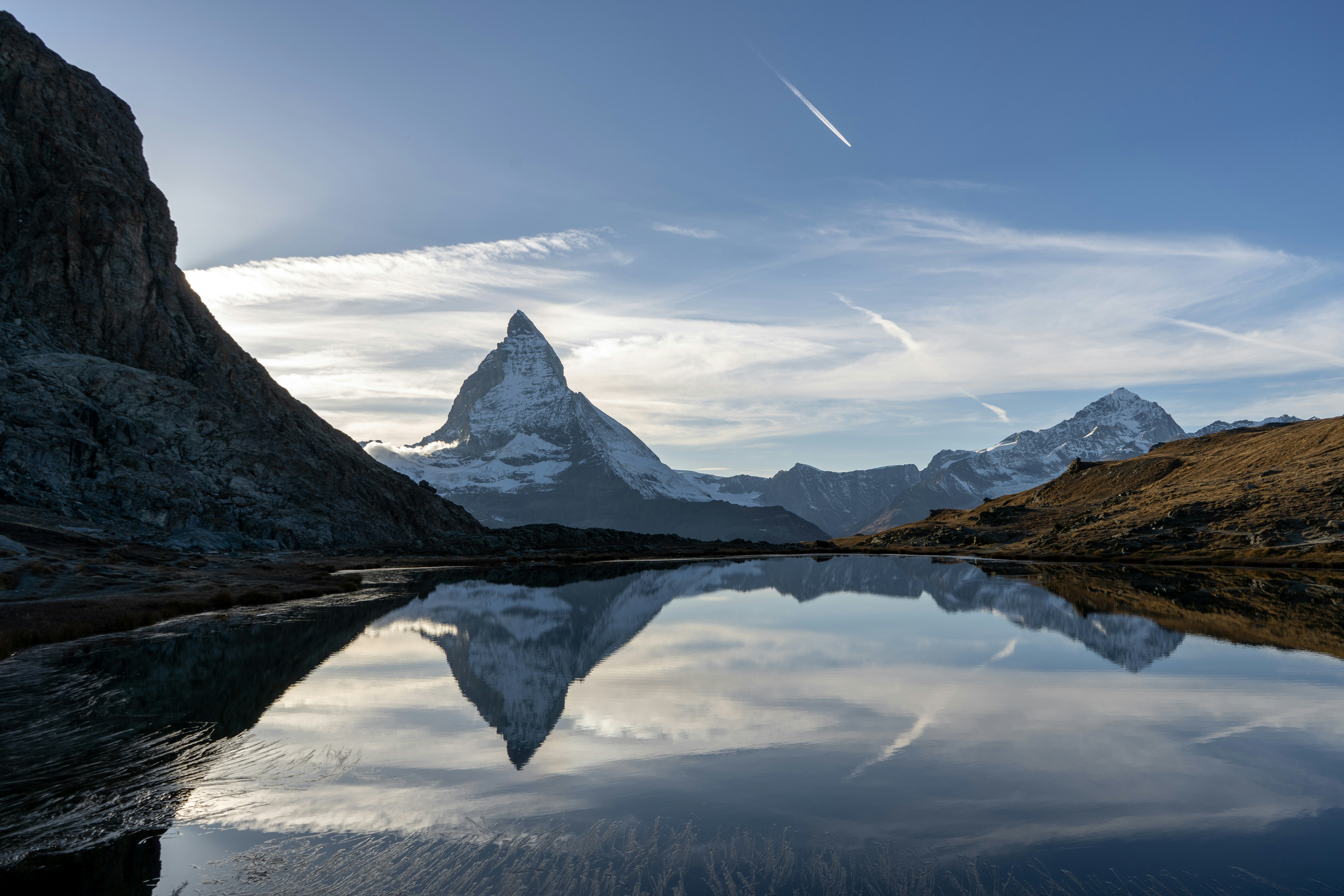 Matterhorn-fjellet med snødekt topp reflektert i en rolig innsjø under en klar blå himmel med skyer, omgitt av røft terreng