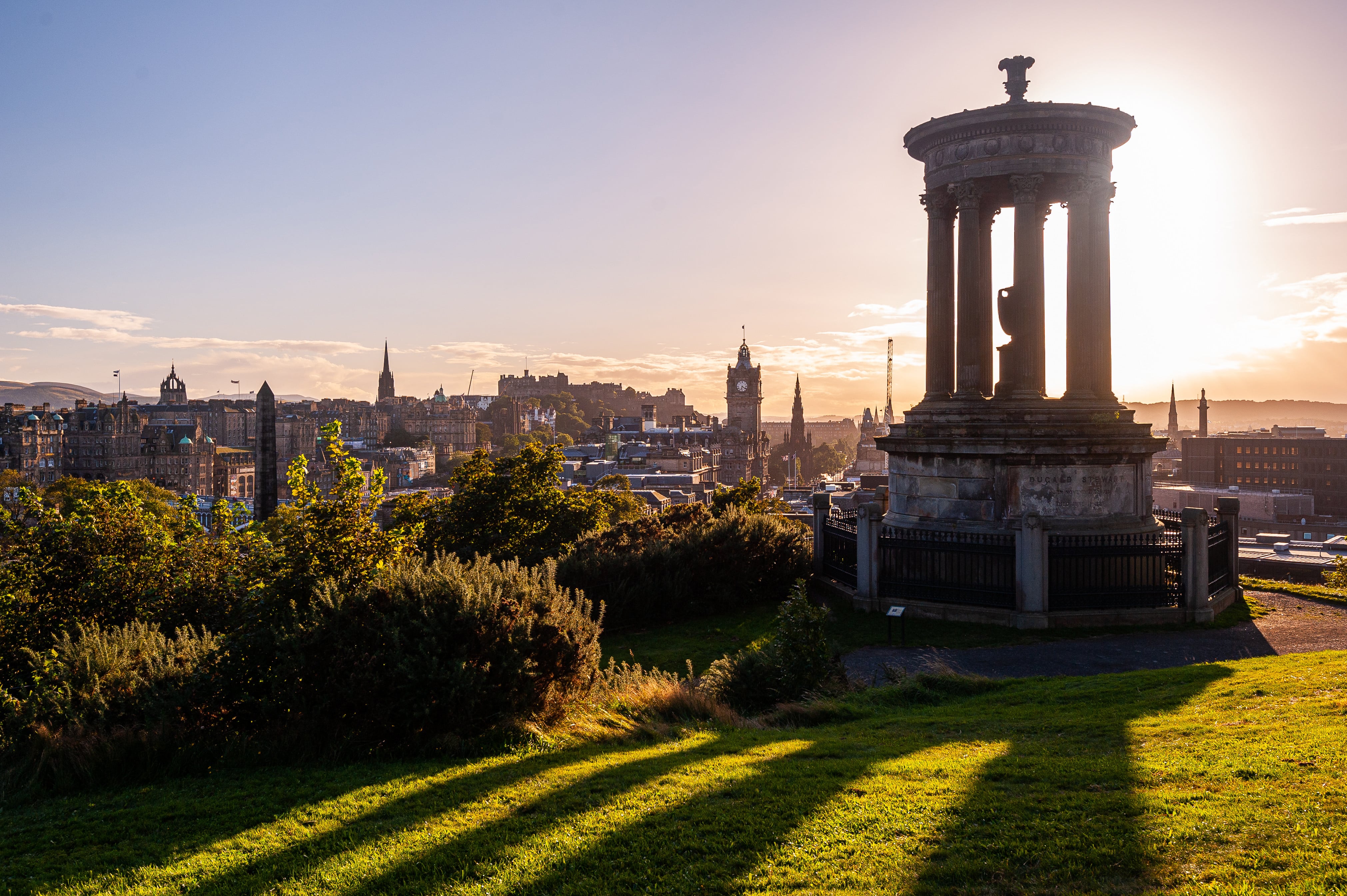 Sunset view of the Dugald Stewart Monument on Calton Hill overlooking Edinburgh, Scotland, with the city skyline and historic architecture in the background.