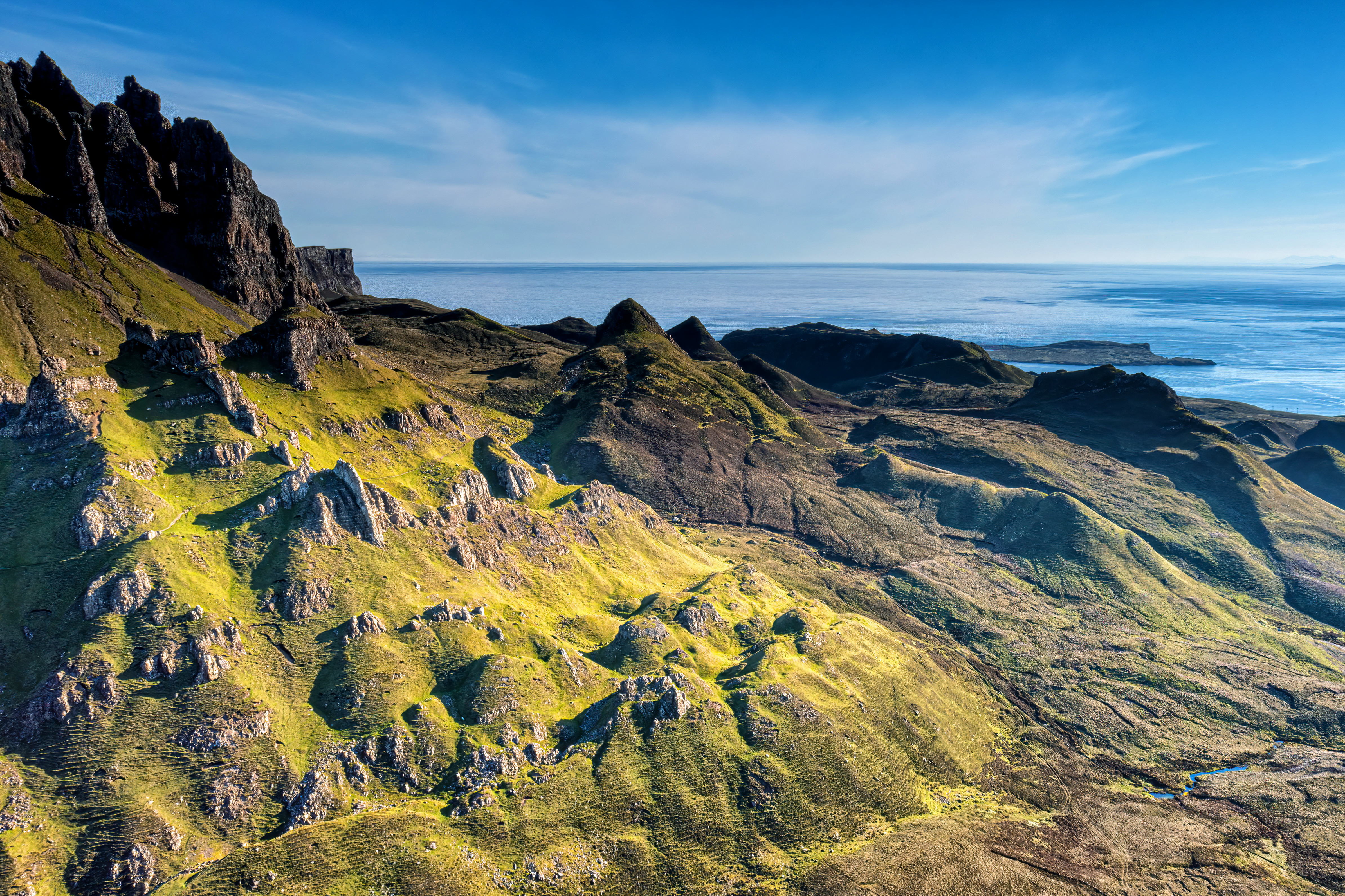 Scenic aerial view of the Quiraing landslip on the Isle of Skye in Scotland, featuring rolling hills with vibrant green grass under a clear blue sky, overlooking the ocean