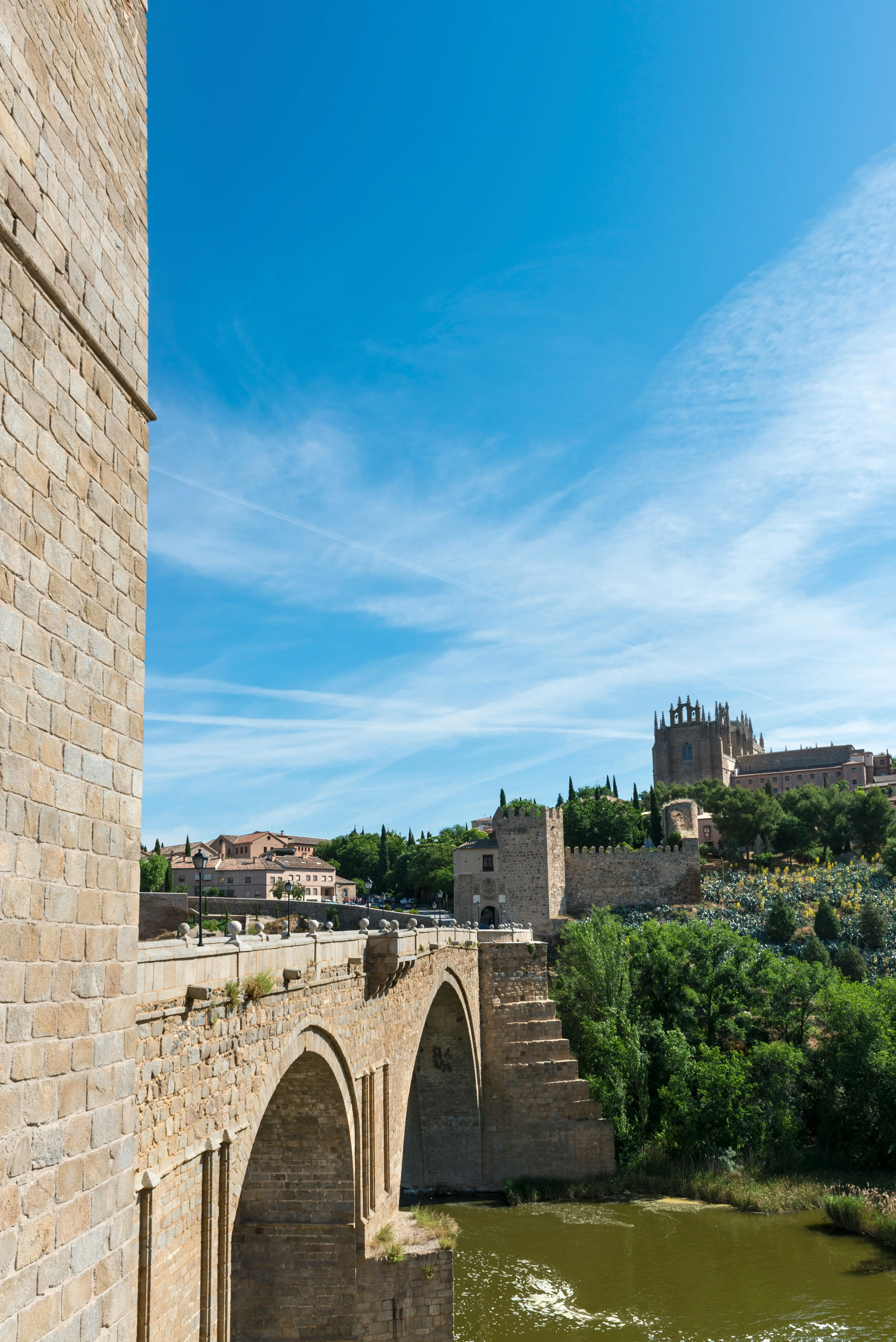 Historic stone bridge over river in Toledo, Spain, with medieval architecture and lush greenery under a clear blue sky