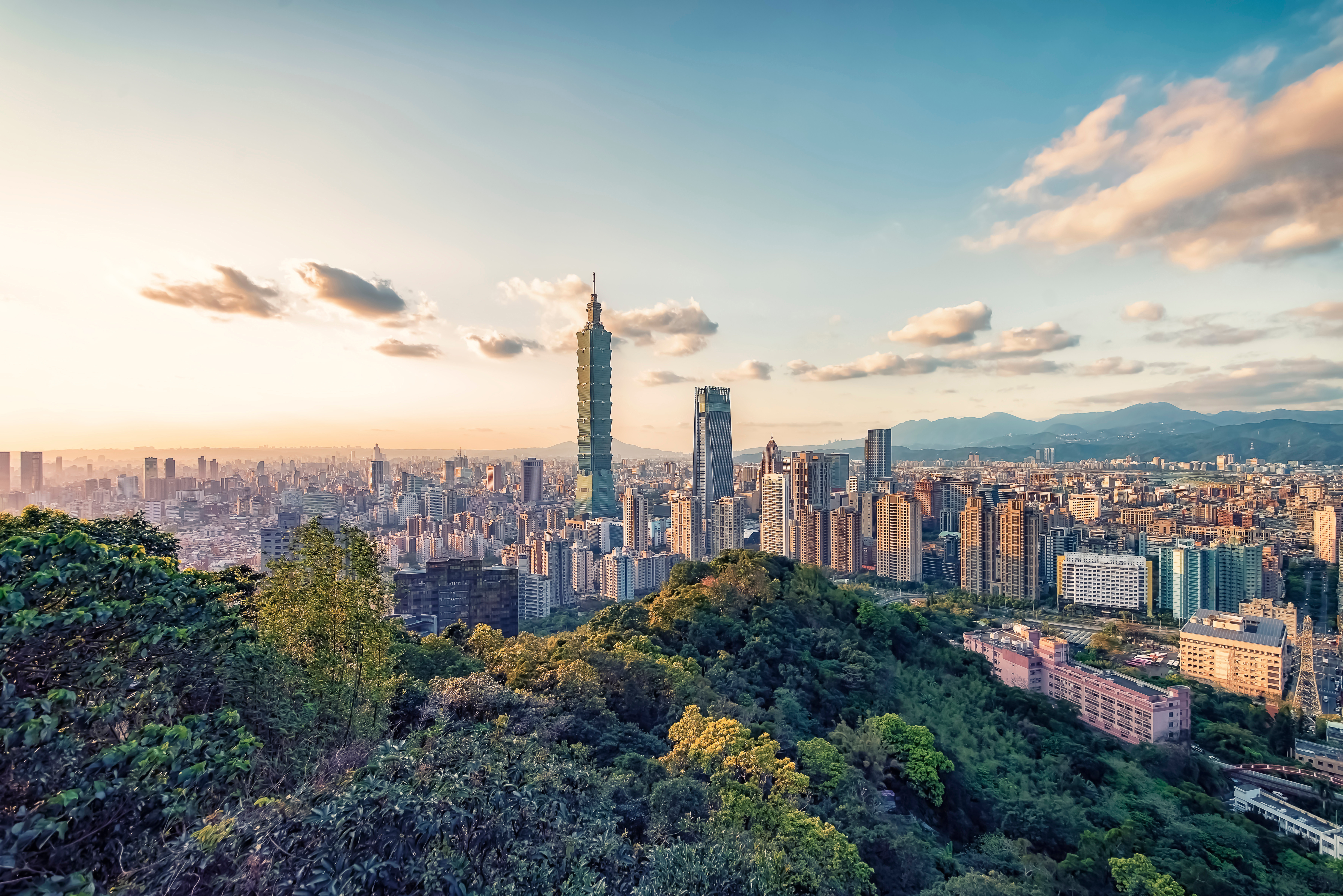Skyline of Taipei, Taiwan, featuring Taipei 101, with lush green hills in the foreground and a vibrant cityscape under a bright, cloudy sky