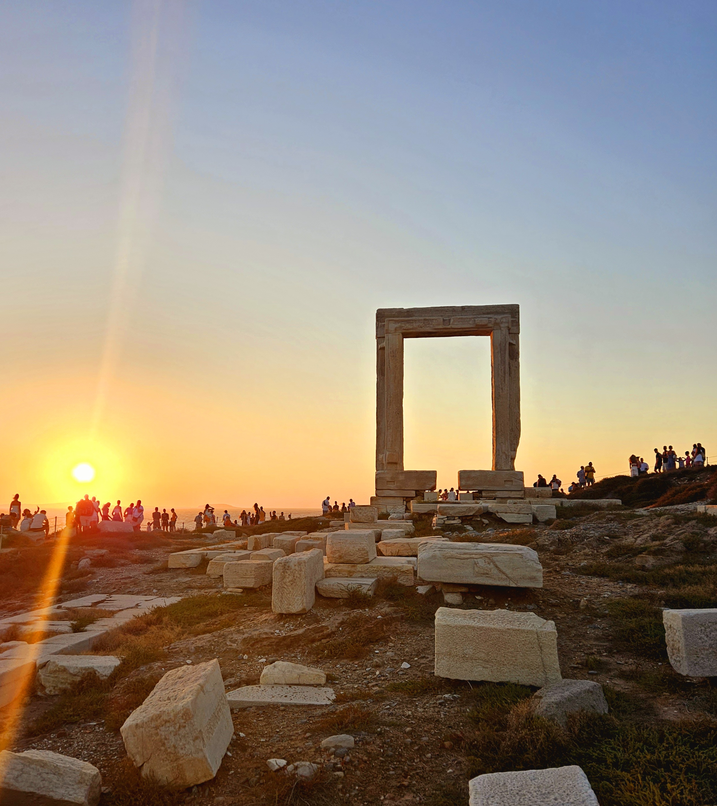 Sunset view of the Portara gate on the Greek island of Naxos, with scattered ancient stone ruins in the foreground.