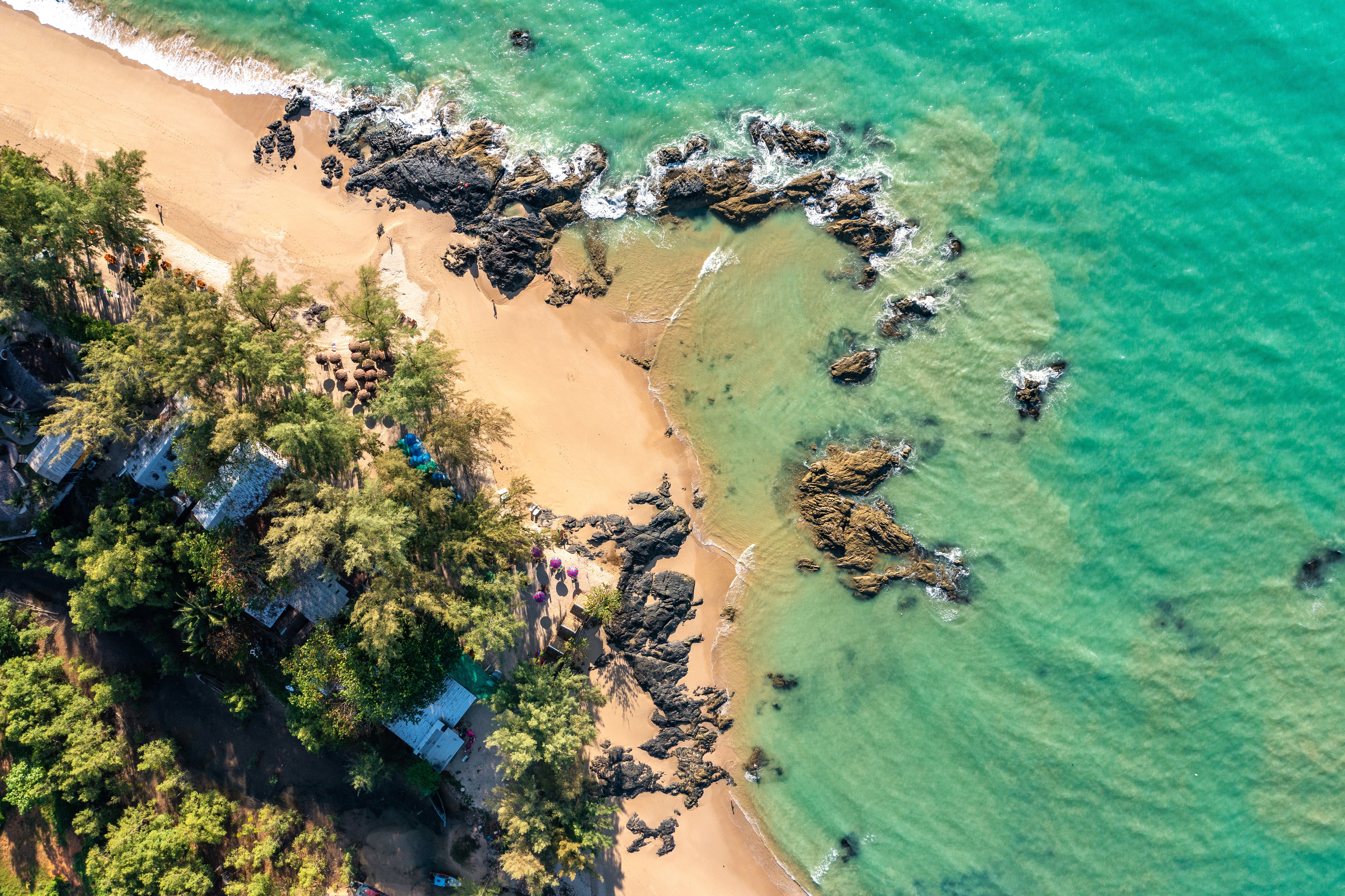 Aerial view of a tropical beach with turquoise water, rocky formations, and a sandy shore lined with lush green trees. Small huts and umbrellas are visible along the shoreline, offering a serene coastal escape