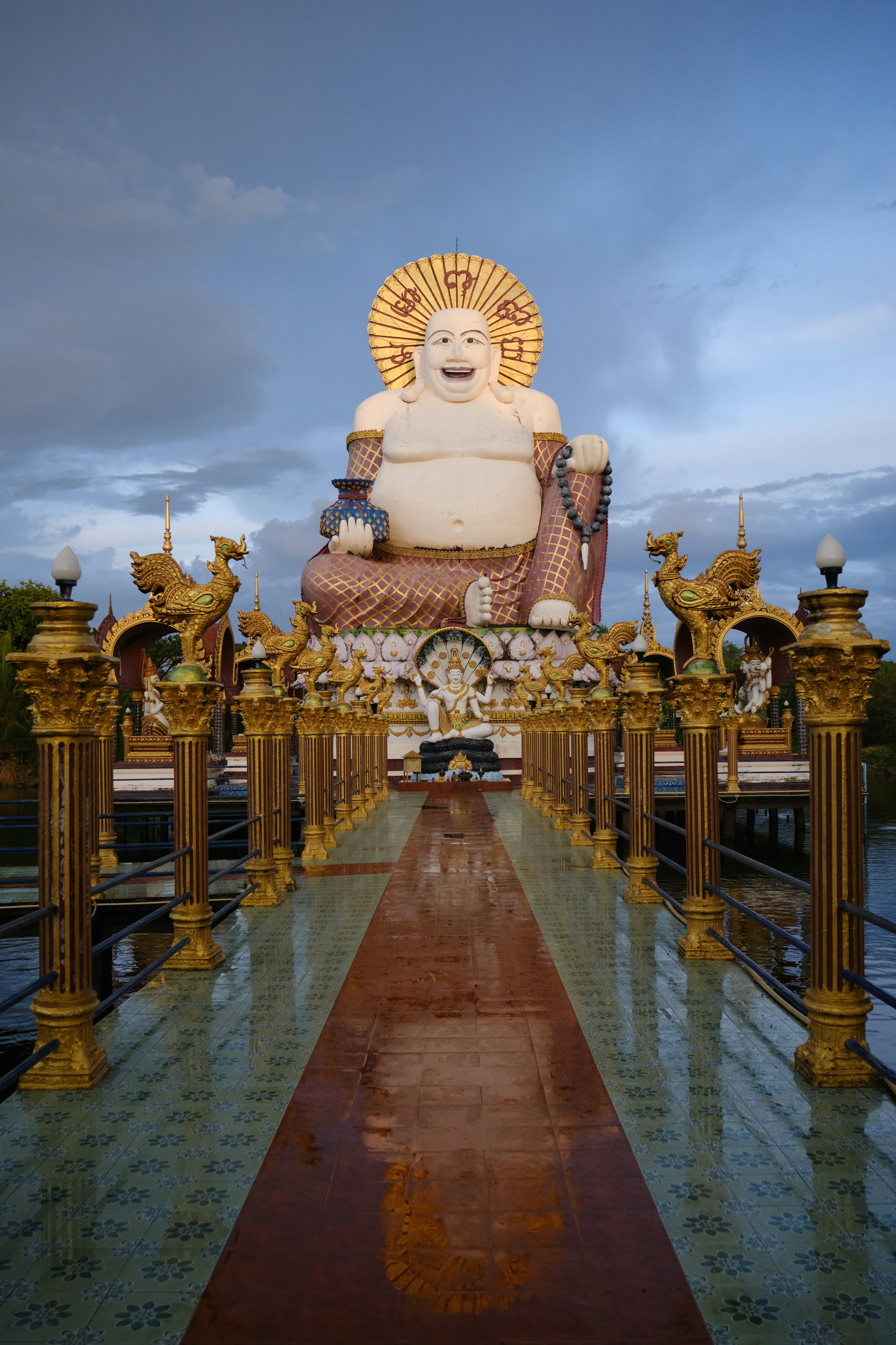Large white Buddha statue with golden decorations at Wat Plai Laem temple in Koh Samui, Thailand, viewed from a decorated walkway with ornate pillars during sunset