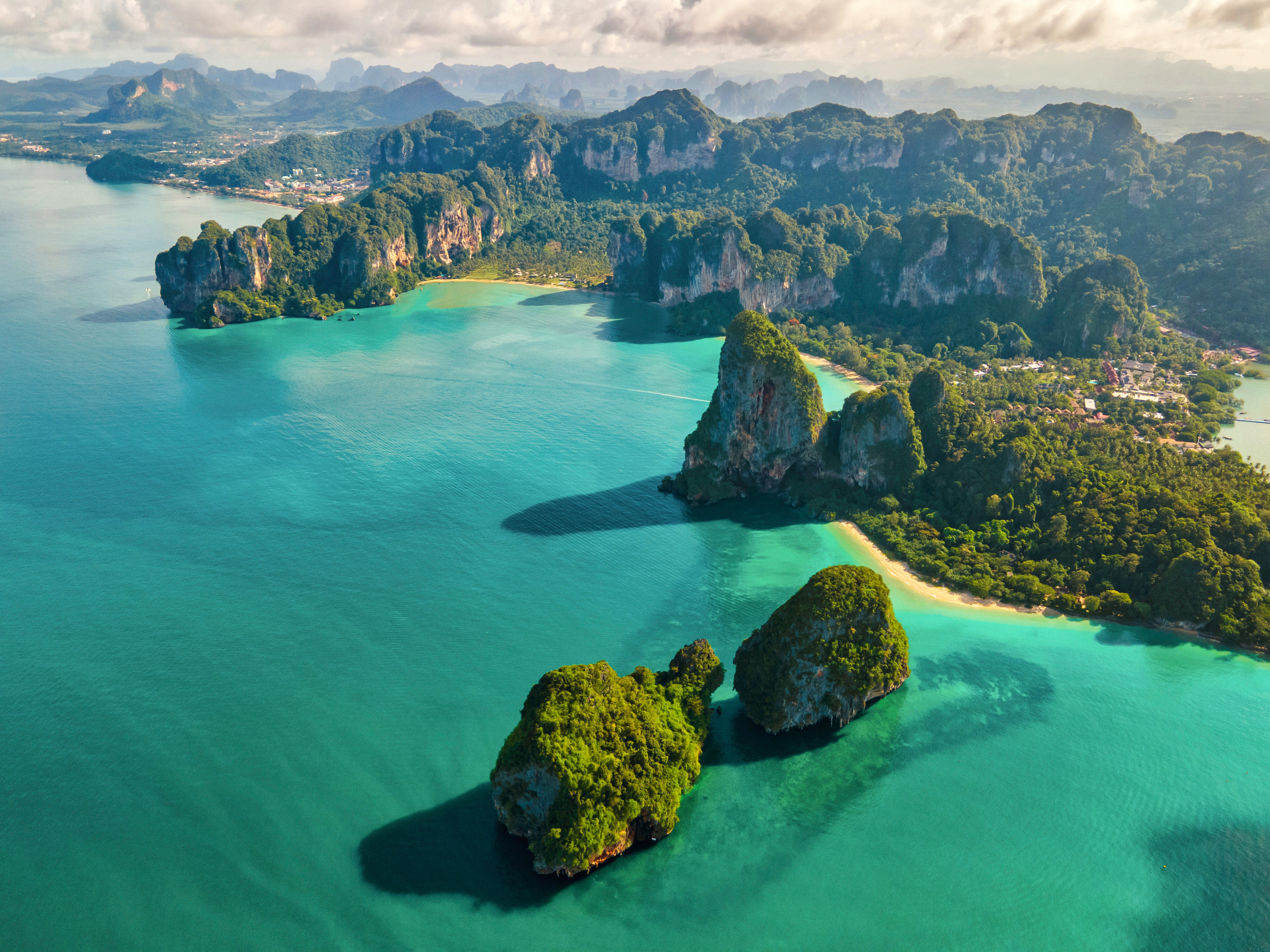 Travel to Krabi - Aerial view of tropical coastline with lush green cliffs, clear turquoise water, and sandy beaches.