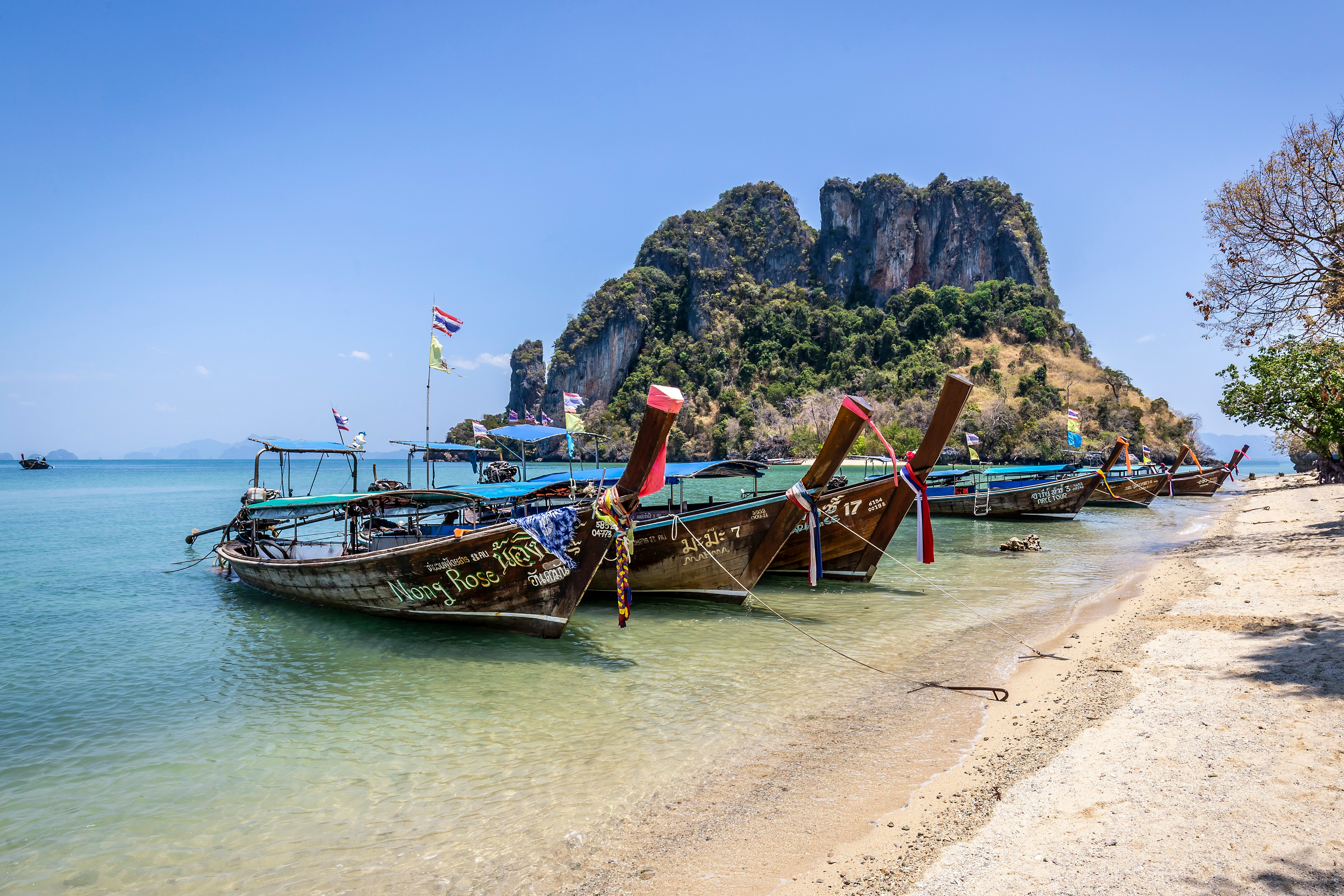 Langhalebåter ved en sandstrand på Krabi, Thailand, foran en kalksteinsklippe under en klar blå himmel