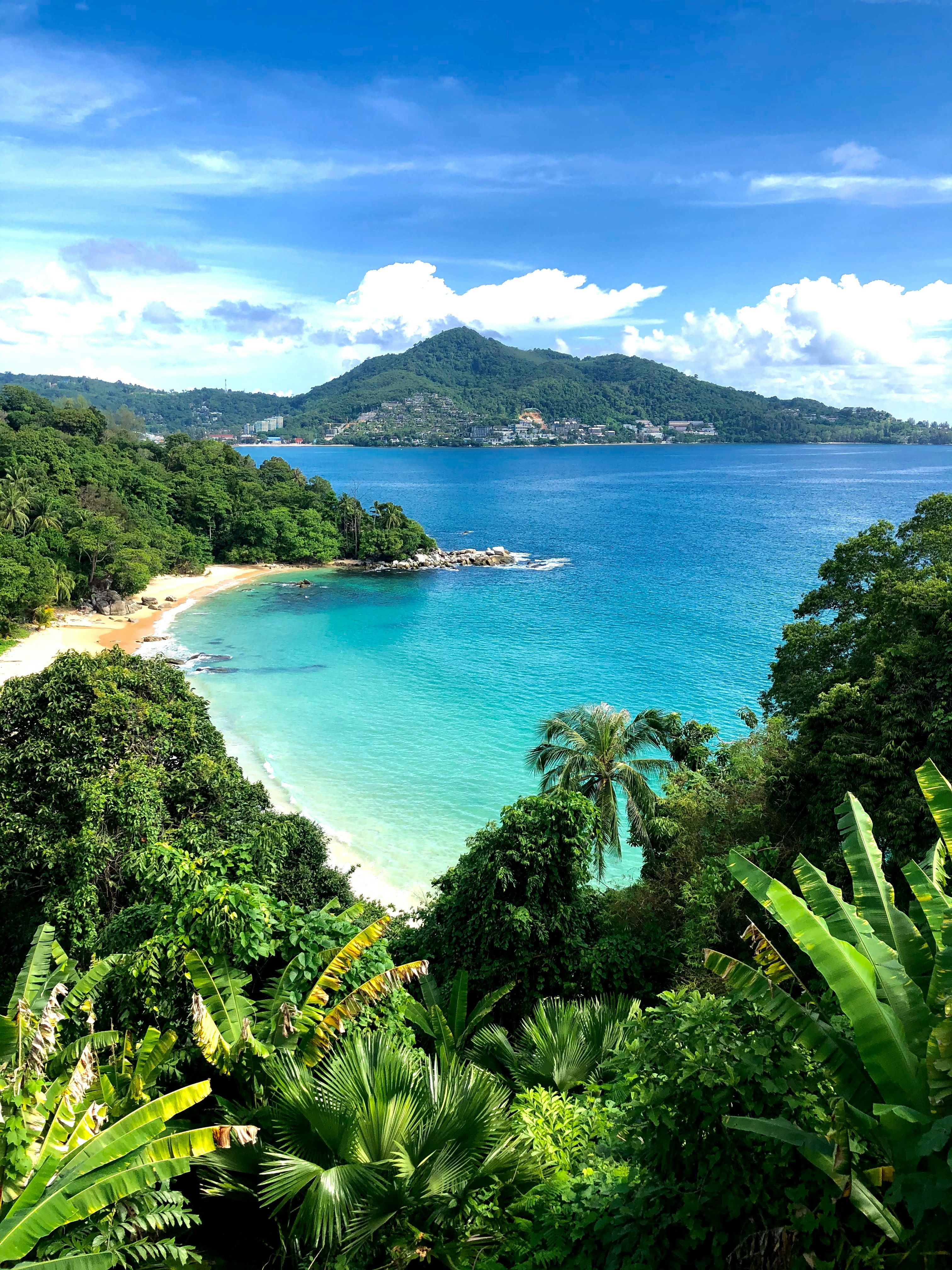 Tropical beach with turquoise water, lush green palm trees, and a mountain in the background under a clear blue sky in Phuket, Thailand