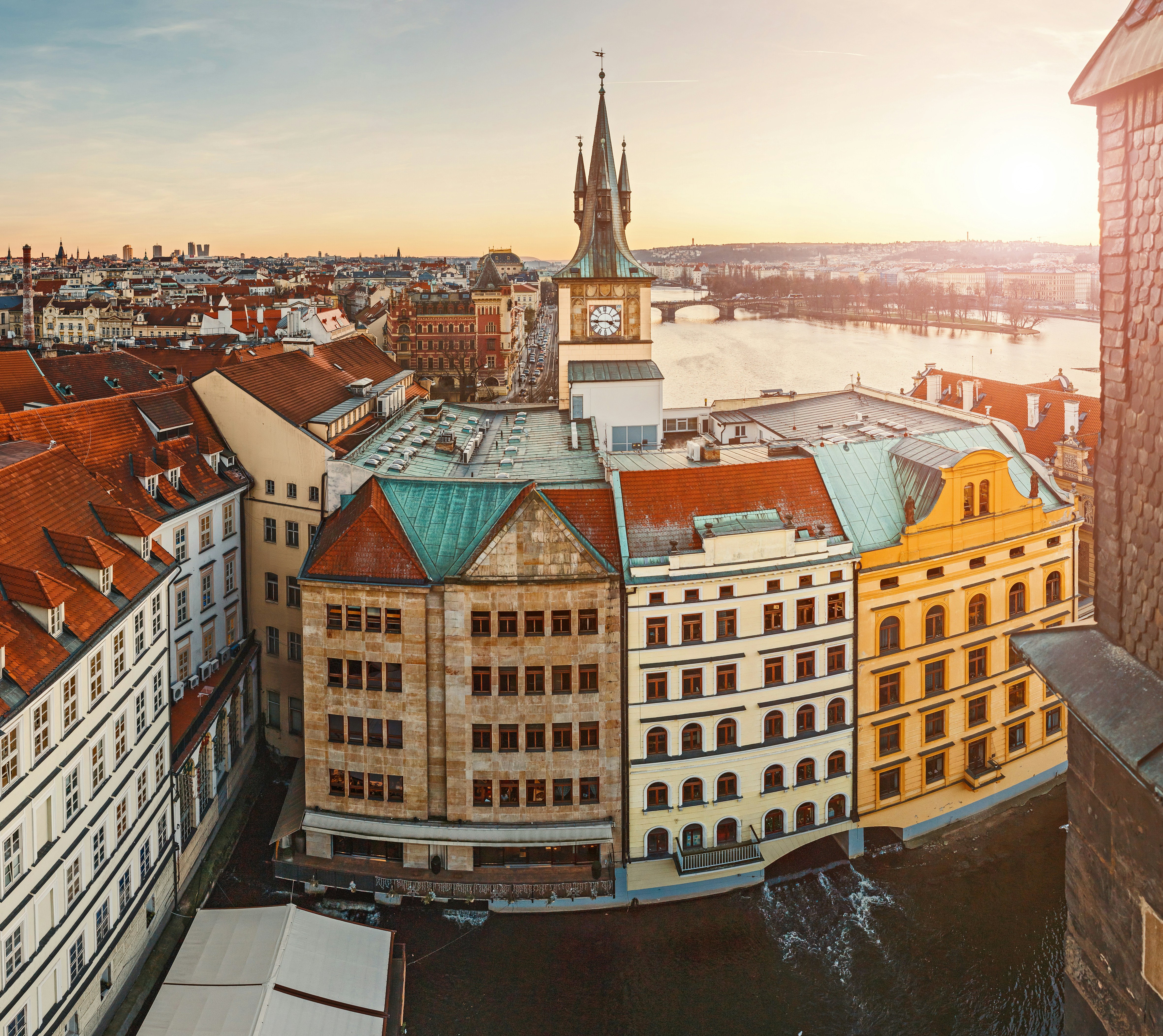 Aerial view of historic buildings in Prague, featuring a clock tower and red-tiled roofs, with the Vltava River and a bright sunset in the background