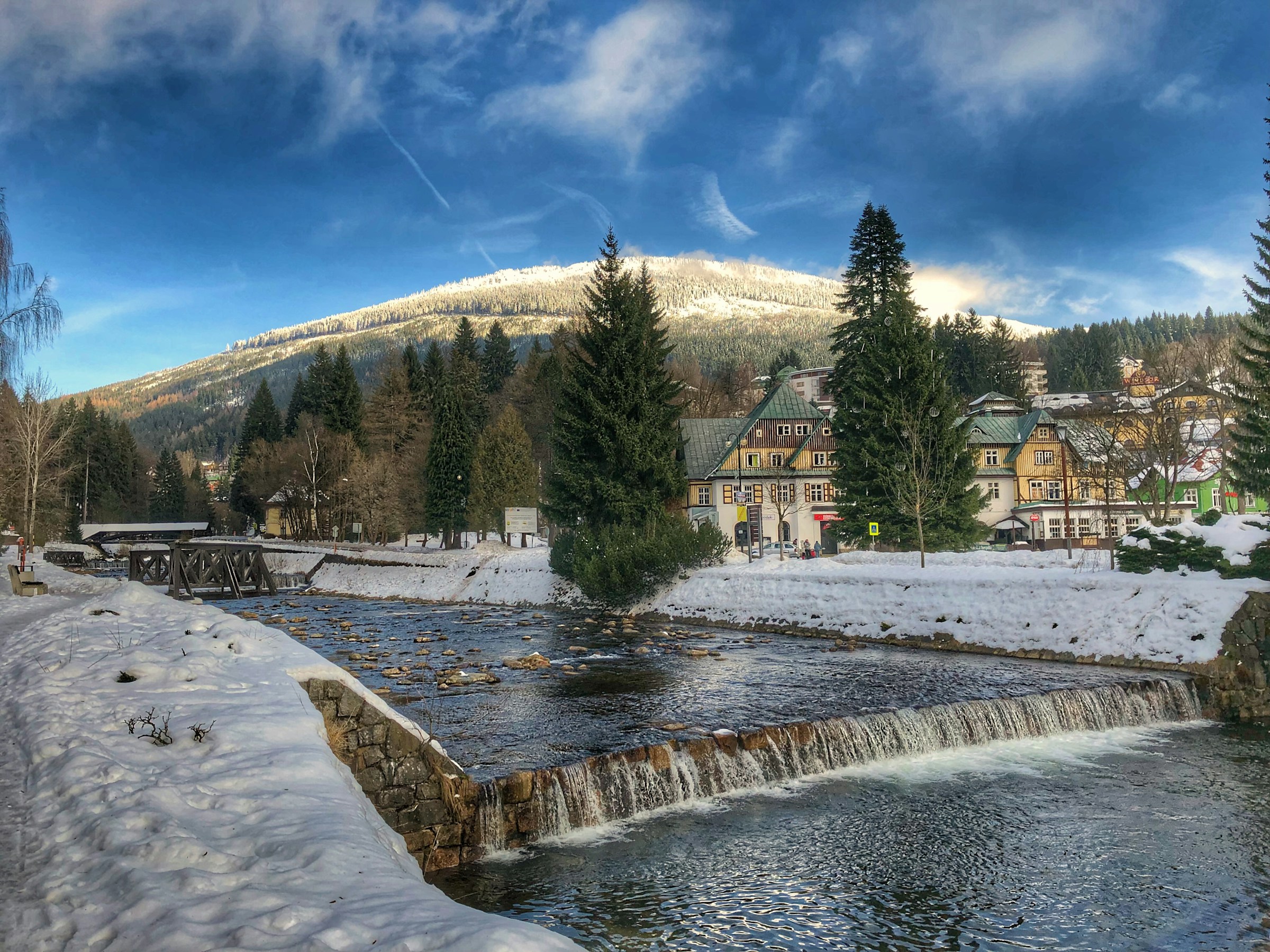 En elv renner gjennom en liten snødekt landsby i Spindleruv Mlyn med fjell og blå himmel i bakgrunnen