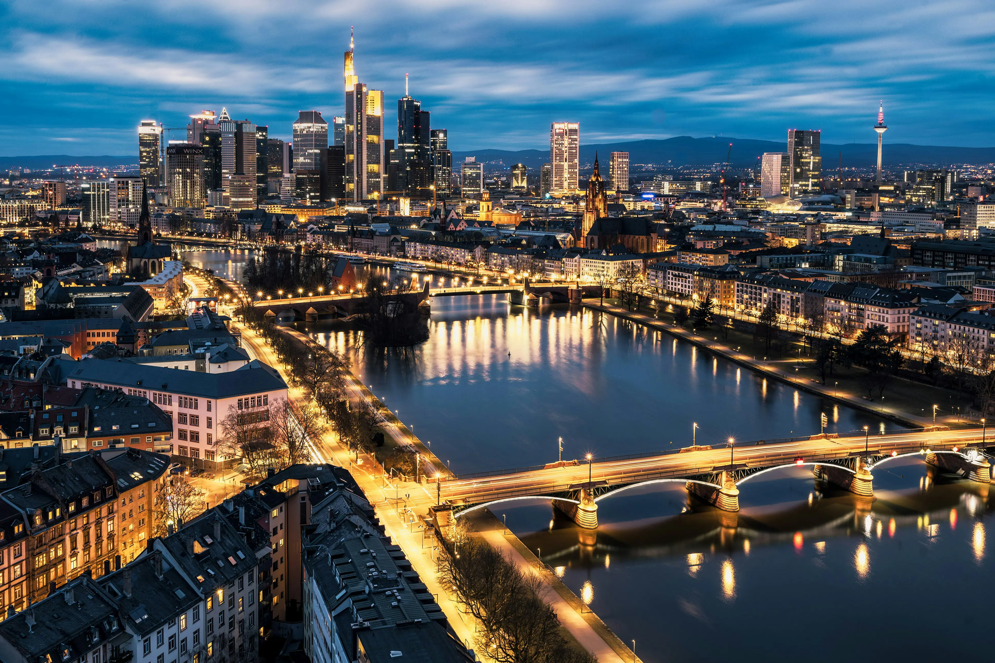 Frankfurts skyline i Tyskland i skumringen med opplyste skyskrapere, som reflekteres i Main-elven og synlige broer, under en blå, skyet himmel