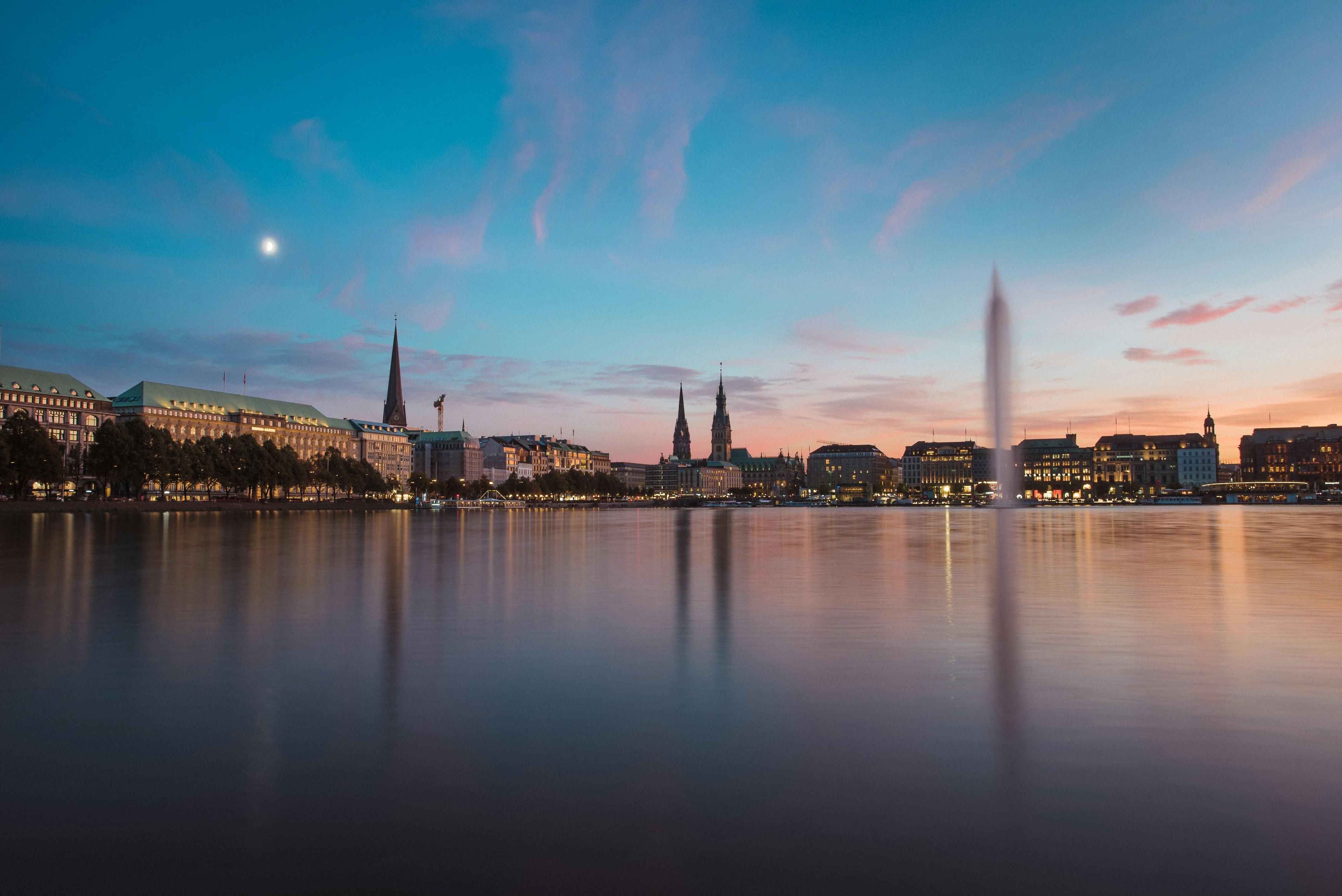 Kveldsutsikt over Alster innsjø i Hamburg med reflekterte lys fra byens skyline og månen synlig på himmelen