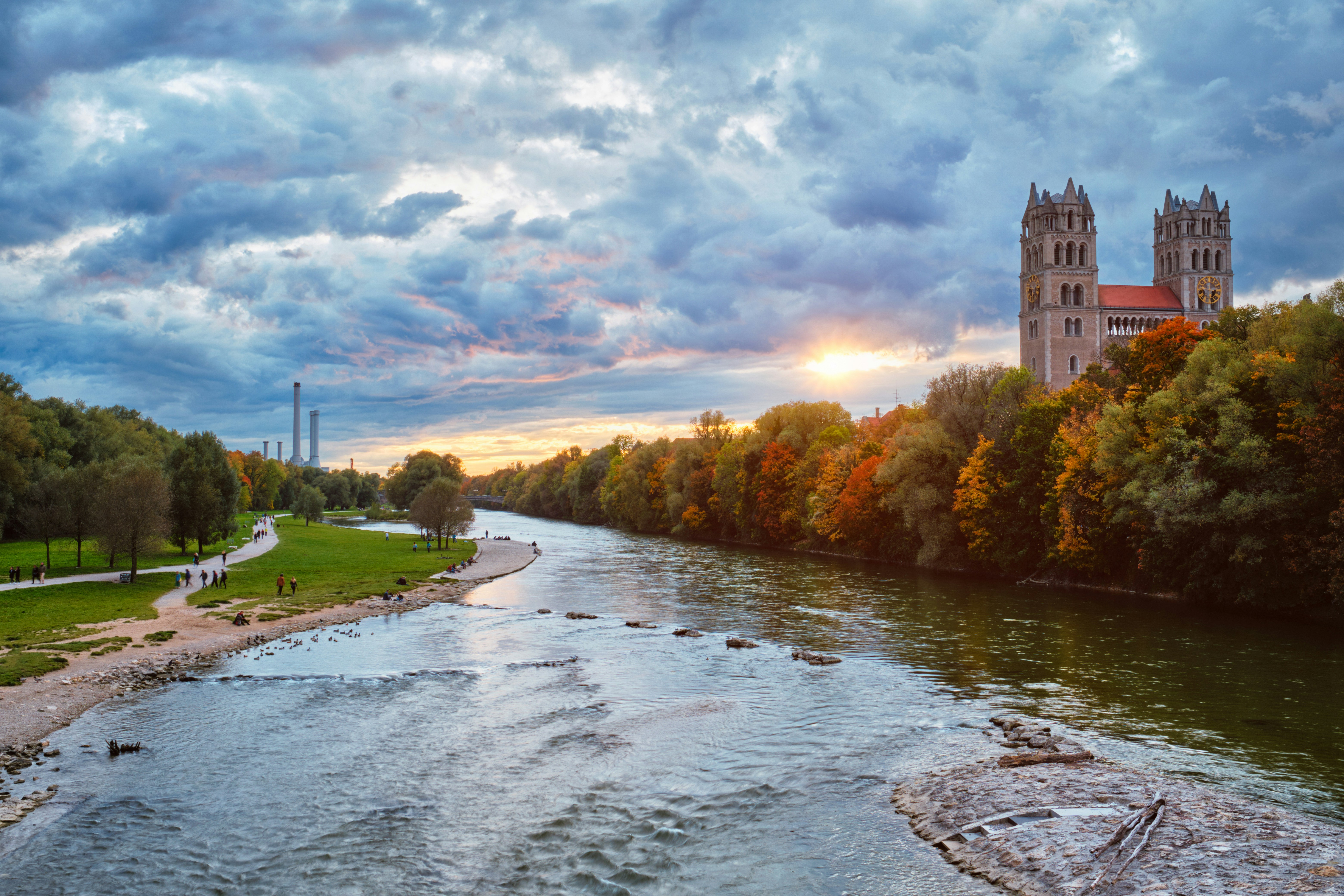 River flowing through a park with a historic building and colorful autumn trees under a dramatic sunset sky