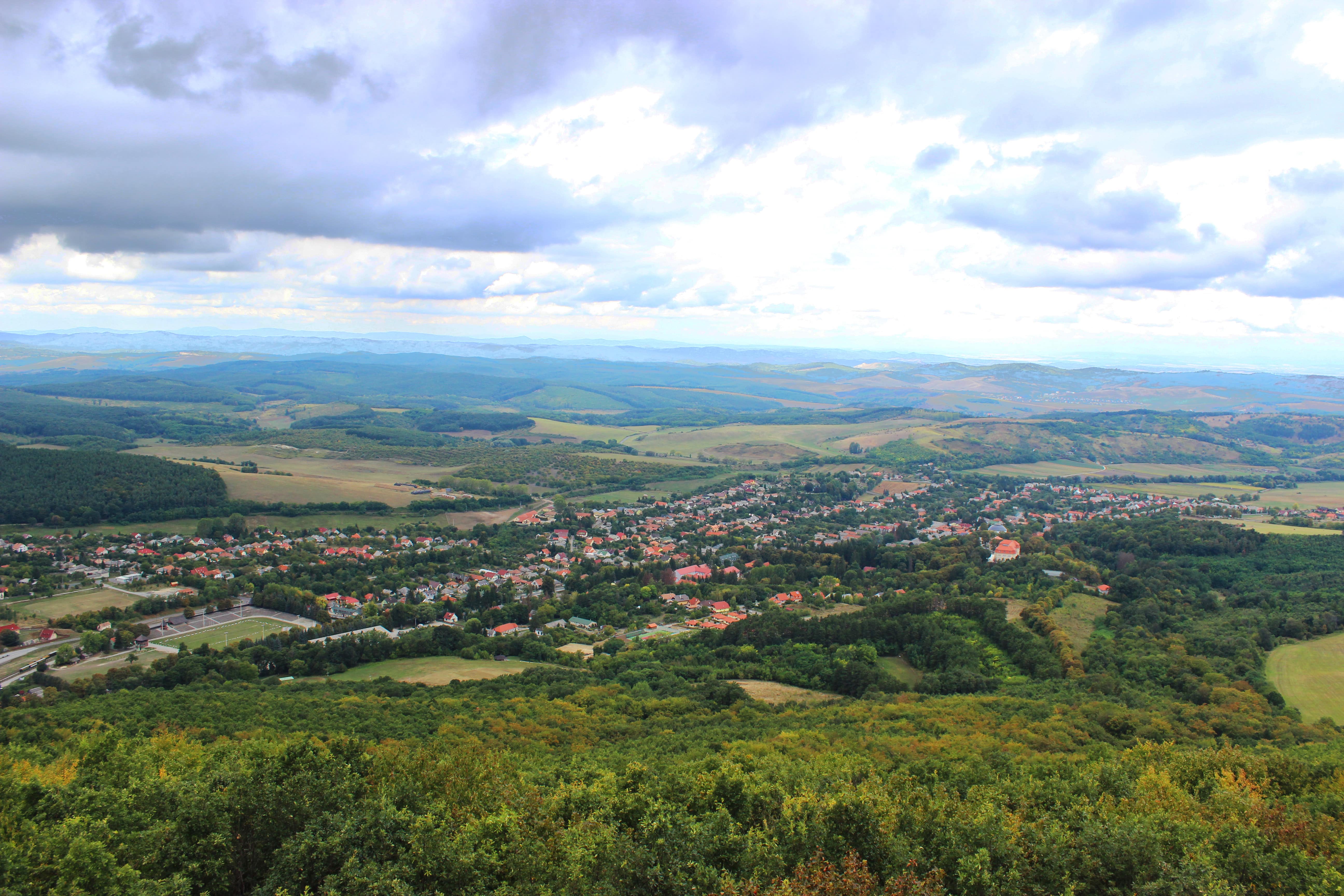 Travel to Hungary - View of a picturesque village nestled in a lush green valley with rolling hills in the background under a cloudy sky.