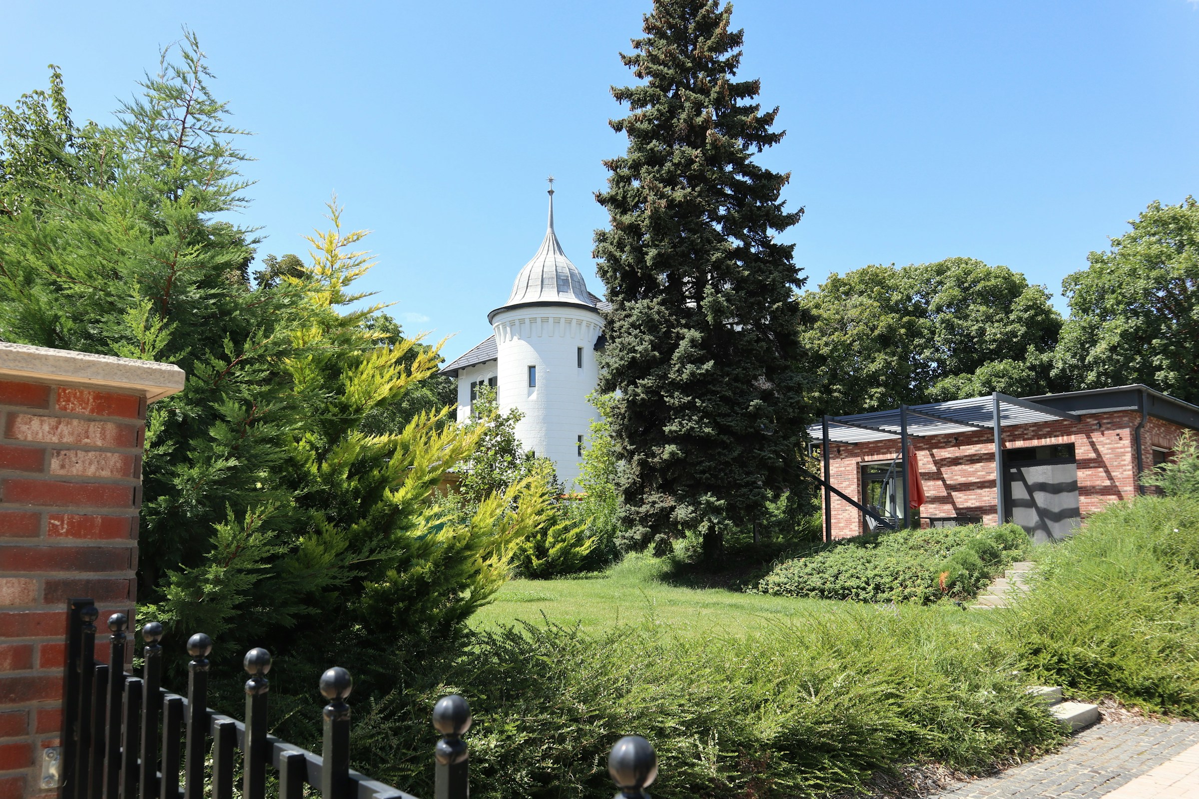 Historic building with a round white tower and conical roof, surrounded by lush greenery and a modern brick structure, under a clear blue sky.