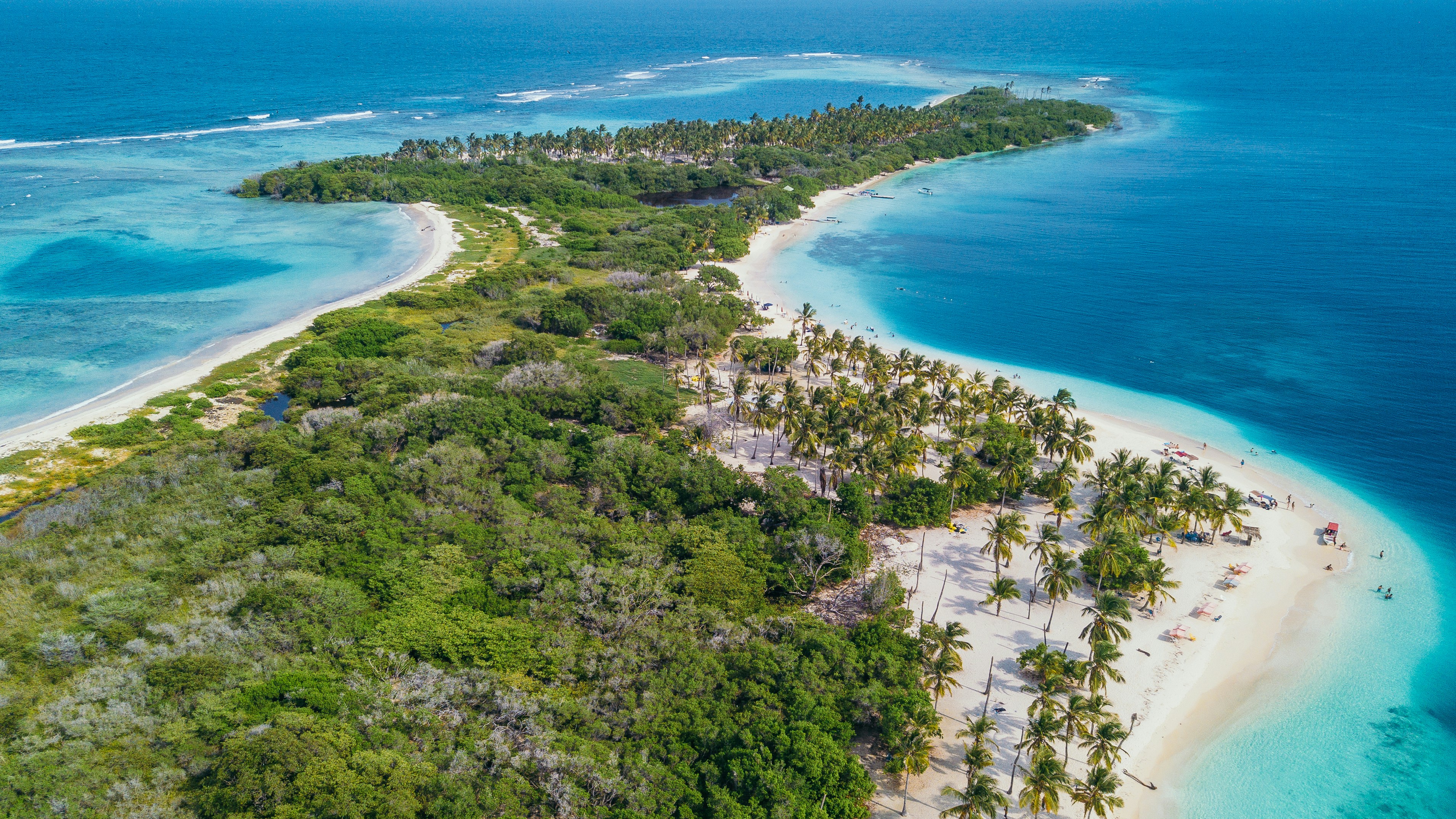 Aerial view of a tropical island with lush greenery, white sandy beaches, and turquoise ocean waters. Palm trees and lounge chairs are visible along the shoreline, depicting a serene paradise setting ideal for vacation